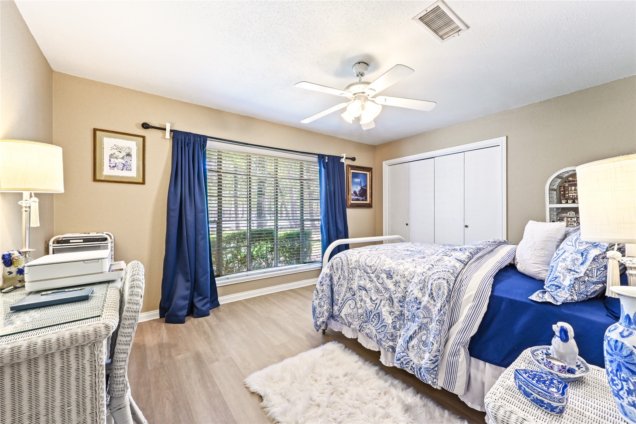 Charming bedroom with natural light through large windows, elegant blue accents, and a cozy atmosphere enhanced by a ceiling fan and soft lighting.