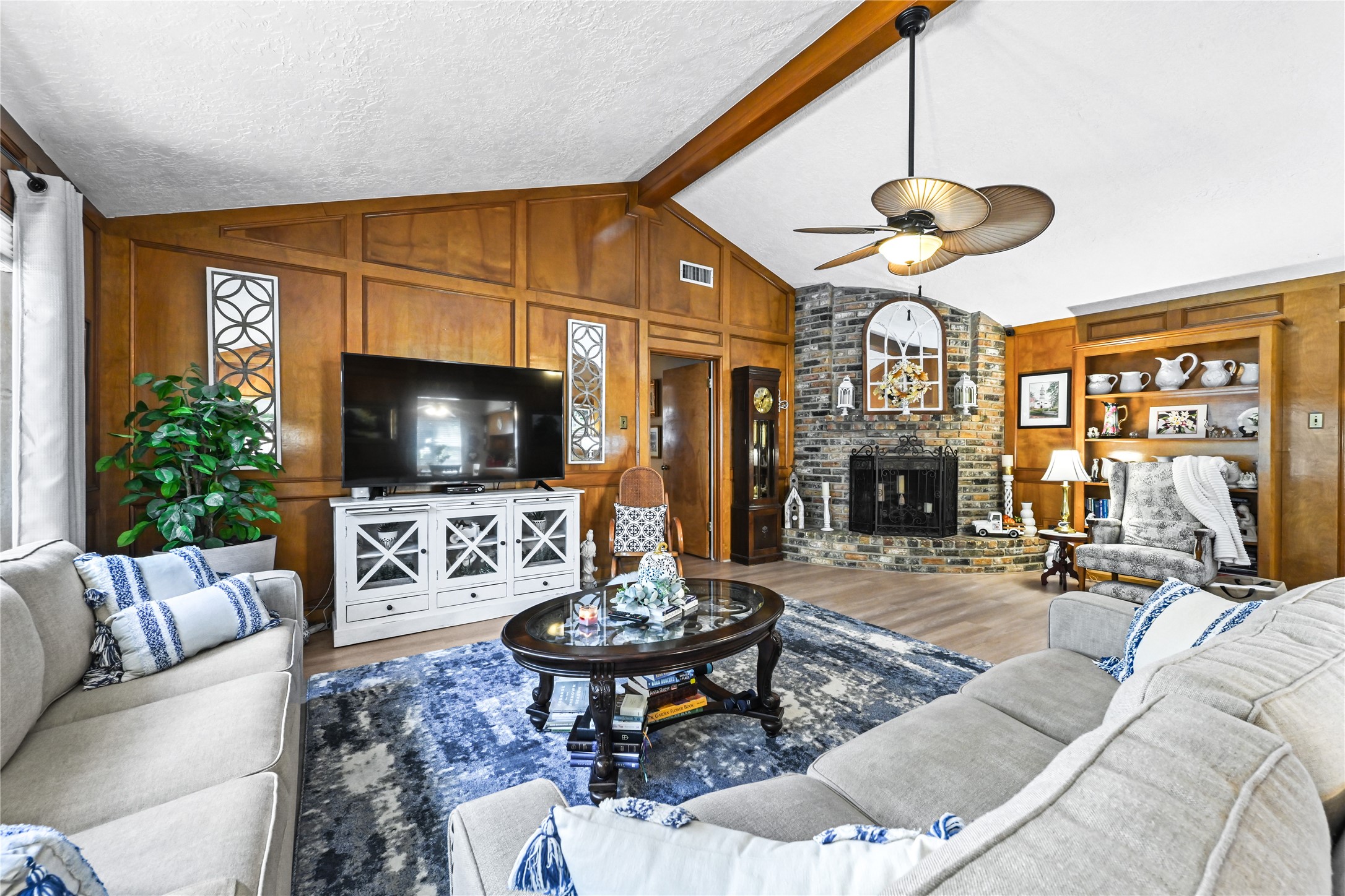 Charming living room featuring a cozy stone fireplace, rich wood paneling, and a vaulted ceiling with an elegant fan. The space is complemented by plush sofas, built-in shelving, and abundant natural light. Charming living room featuring a cozy stone fireplace, rich wood paneling, and a vaulted ceiling with an elegant fan. The space is complemented by plush sofas, built-in shelving, and abundant natural light.