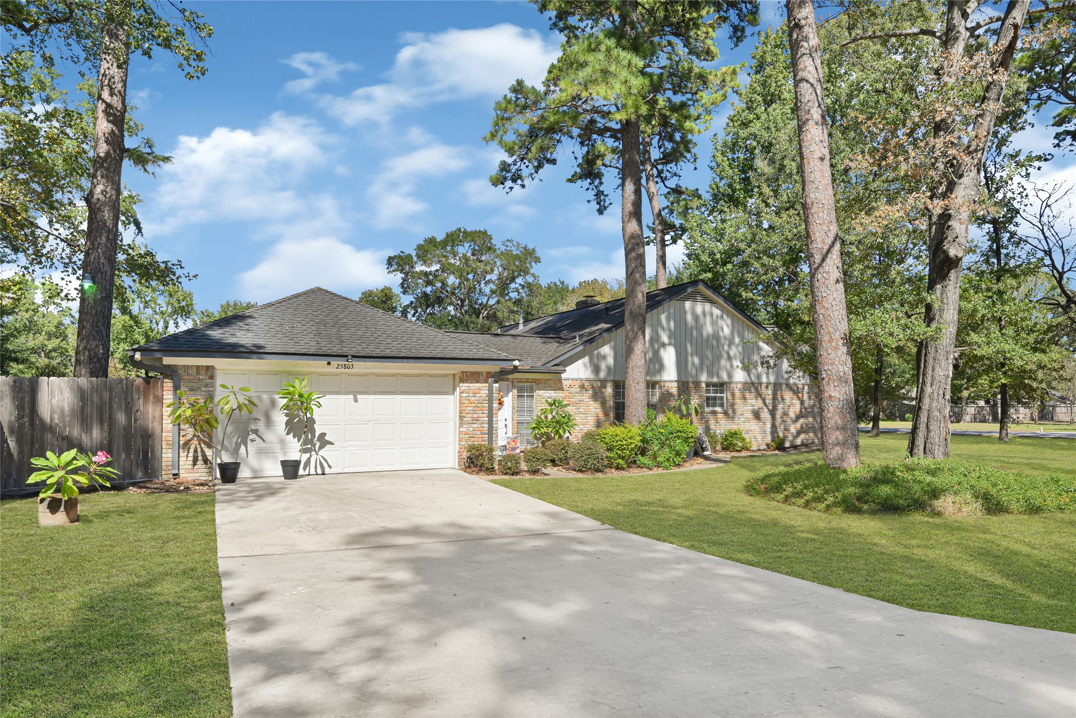 Charming single-story home surrounded by lush trees, featuring a spacious driveway leading to a two-car garage and a beautifully landscaped front yard.