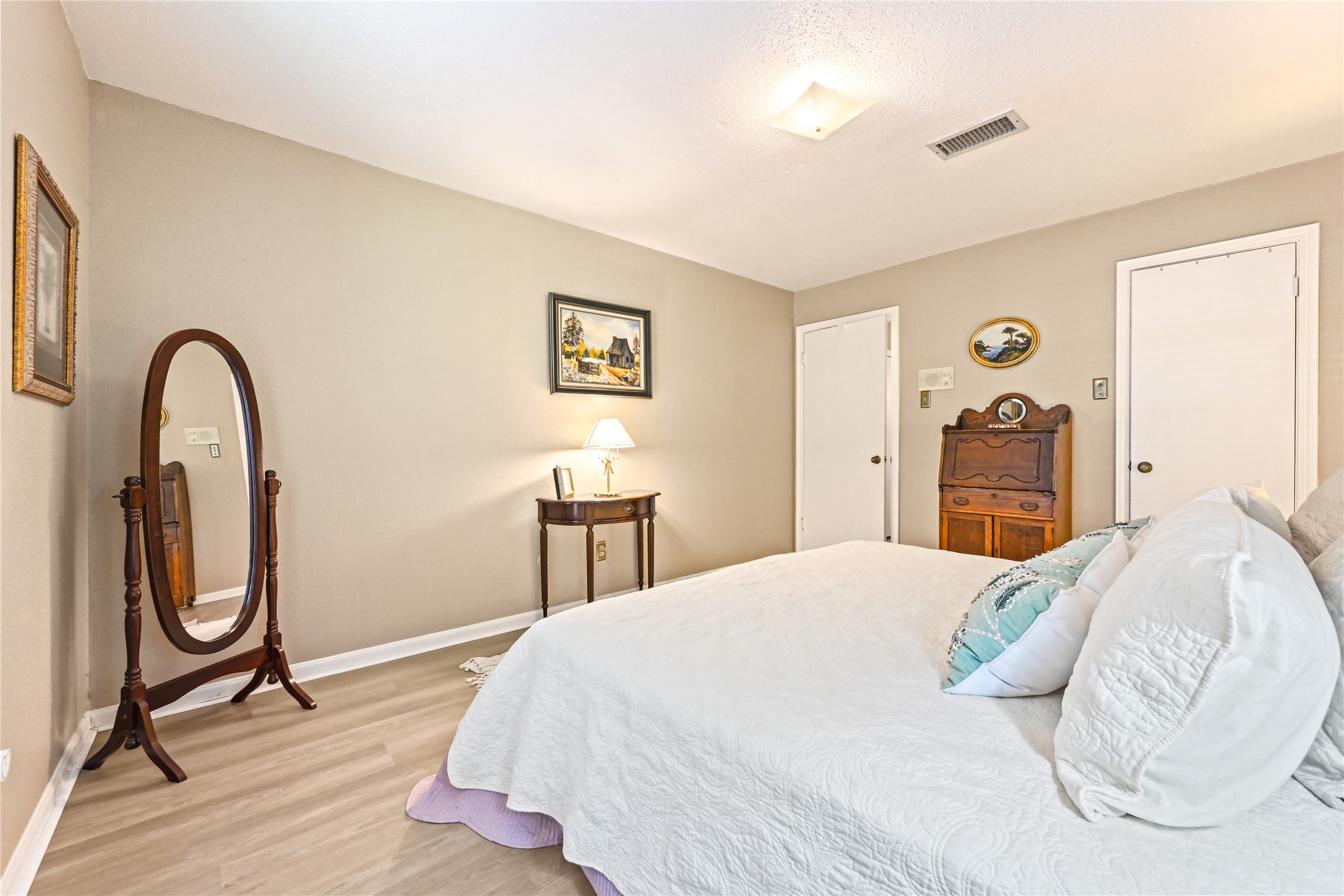 Cozy bedroom with soft neutral walls, featuring a comfortable bed adorned with decorative pillows. A classic floor mirror and elegant wooden furniture add charm, while ample natural light enhances the inviting atmosphere.