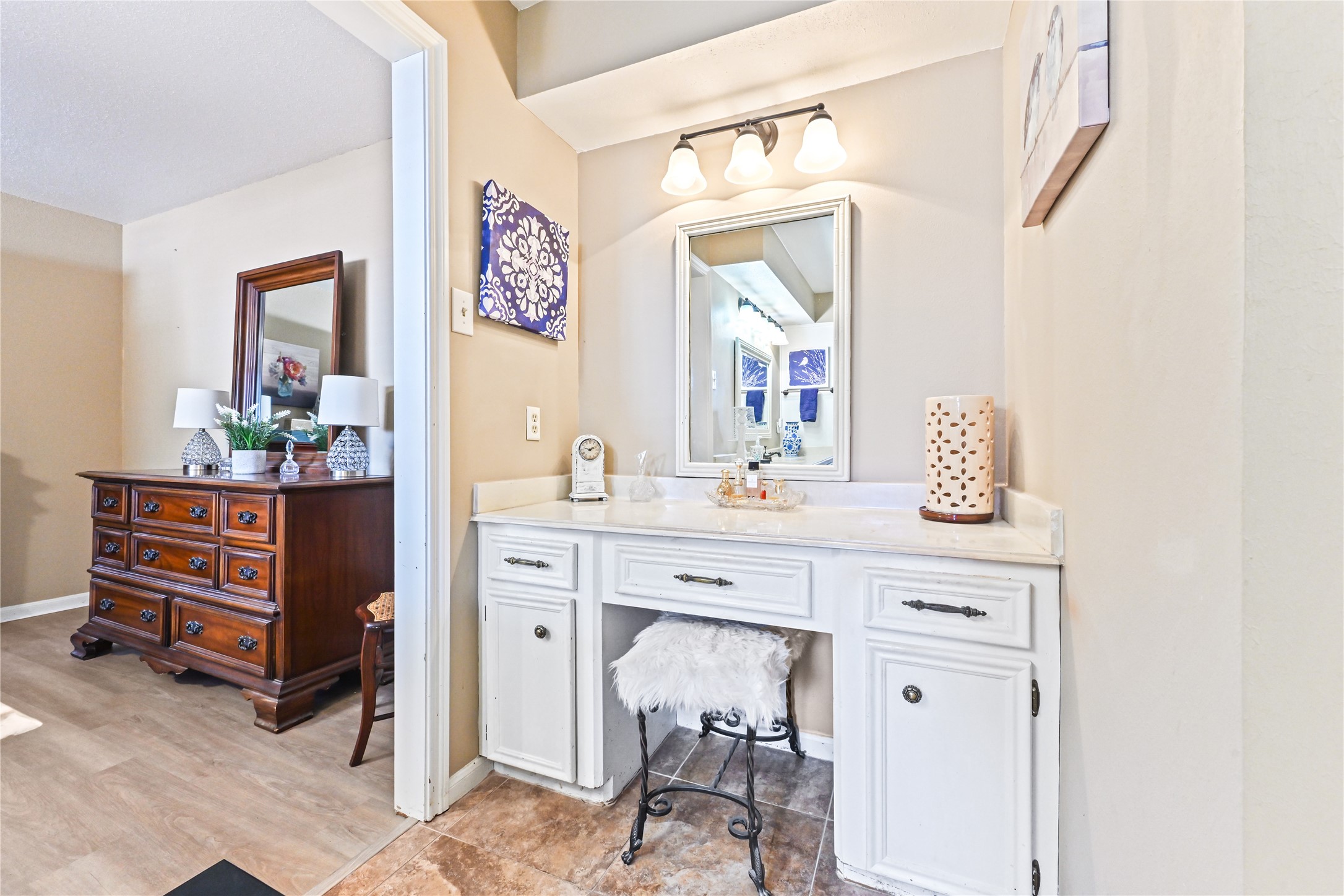 Charming vanity area with a well-lit mirror, elegant white cabinetry, and a cozy seating nook, seamlessly adjoining a warmly furnished bedroom.