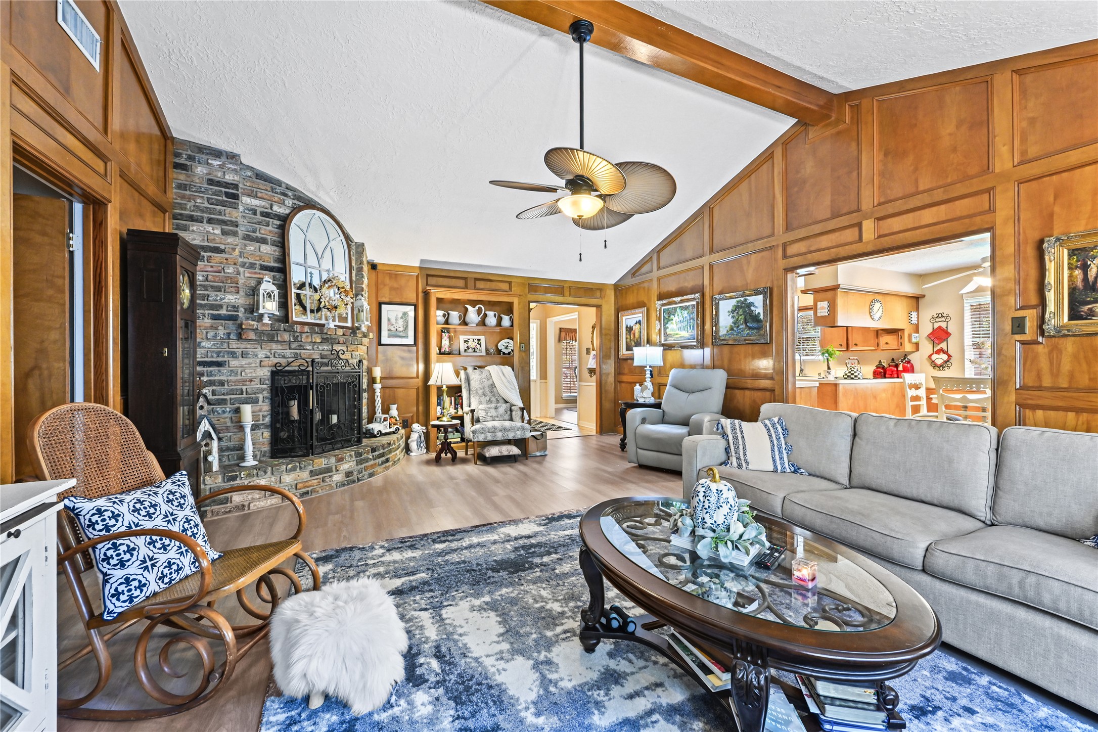 Charming living room featuring vaulted ceilings and warm wood paneling, highlighted by a cozy brick fireplace. The open layout seamlessly connects to a bright kitchen, perfect for entertaining.