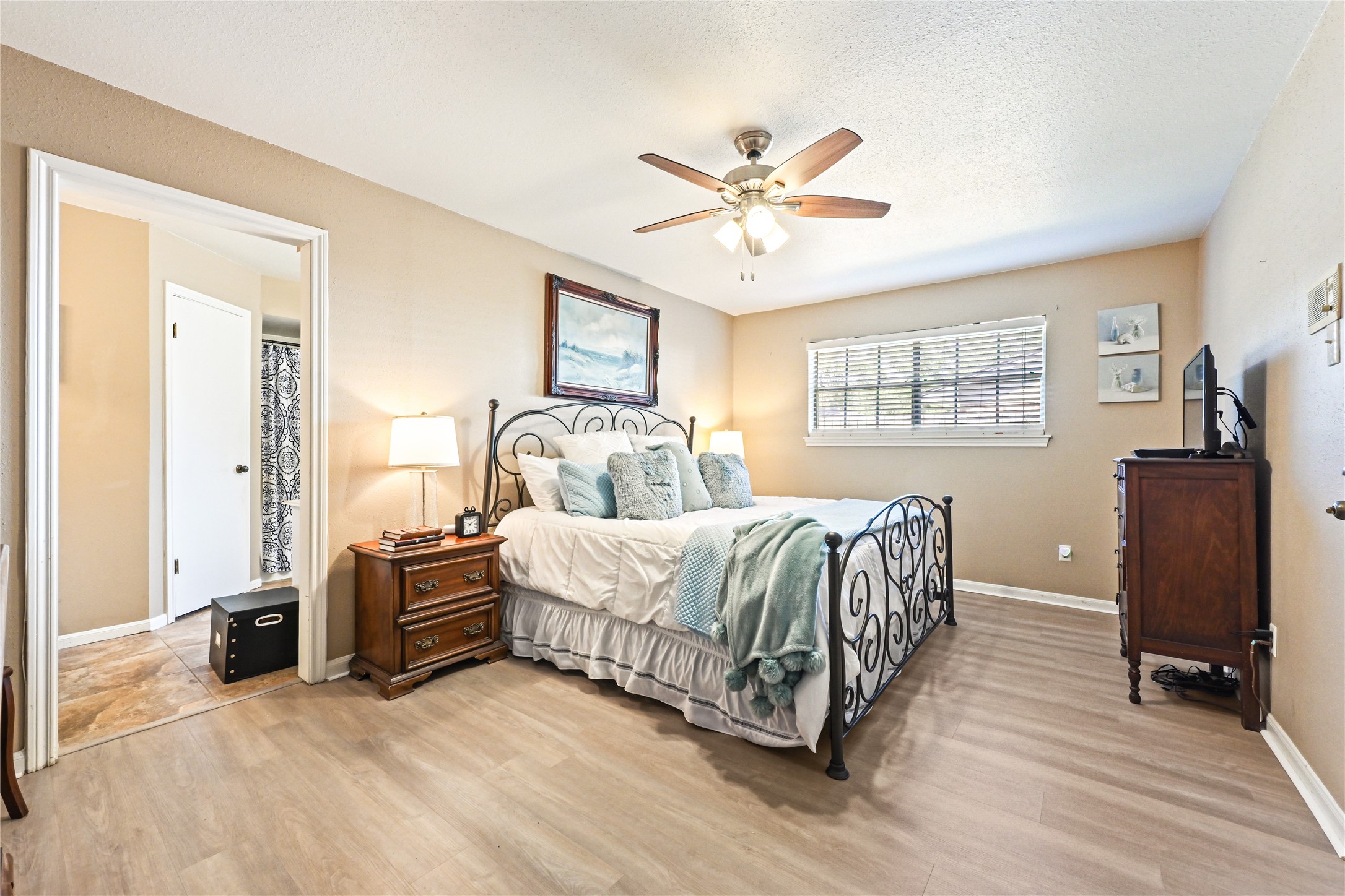 Cozy bedroom featuring a stylish wrought iron bed, warm laminate flooring, and ample natural light from a large window, complemented by a ceiling fan for added comfort. Cozy bedroom featuring a stylish wrought iron bed, warm laminate flooring, and ample natural light from a large window, complemented by a ceiling fan for added comfort.
