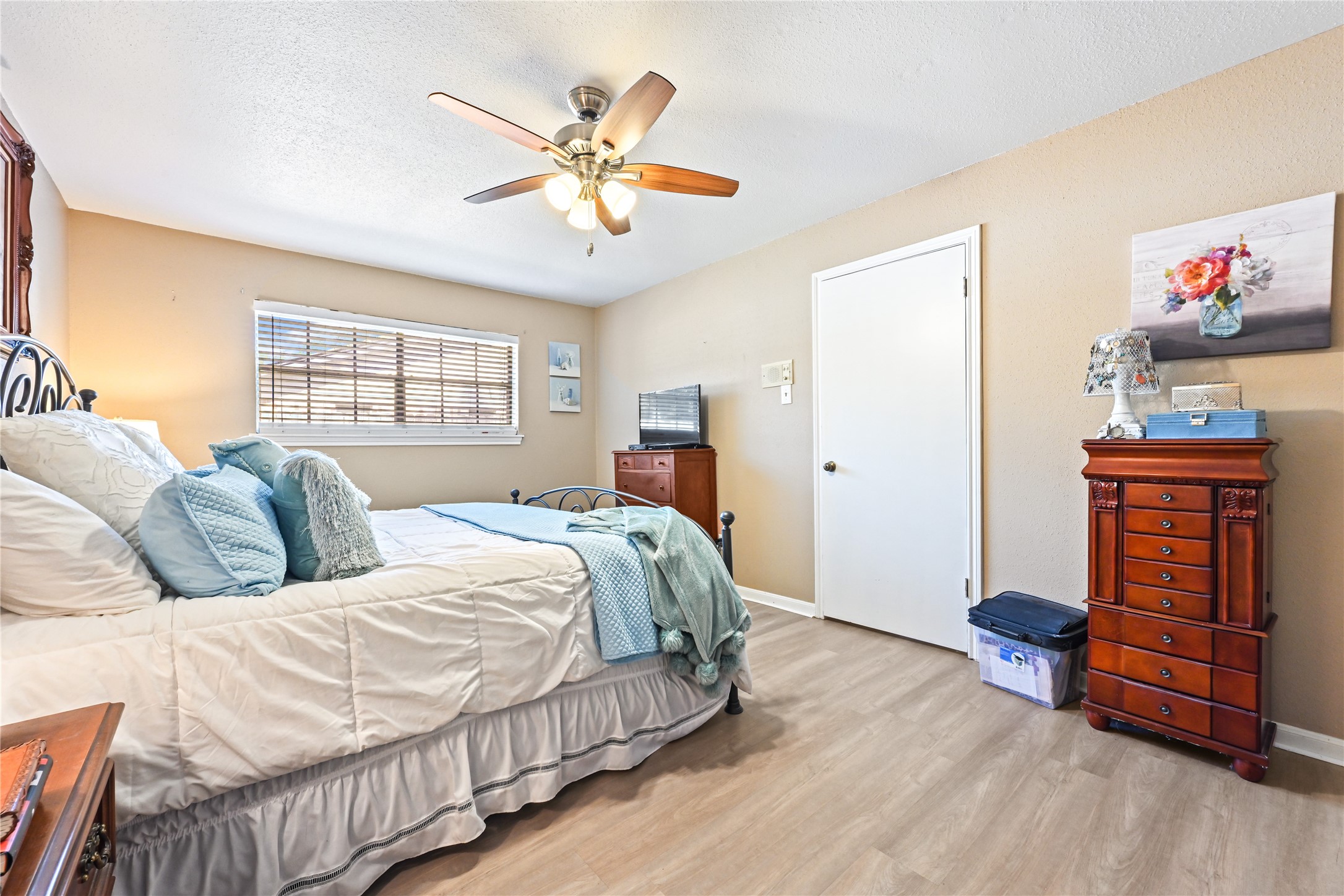 Spacious bedroom with warm, neutral tones features a cozy bed with elegant iron headboard, a ceiling fan for comfort, and ample natural light from a large window.