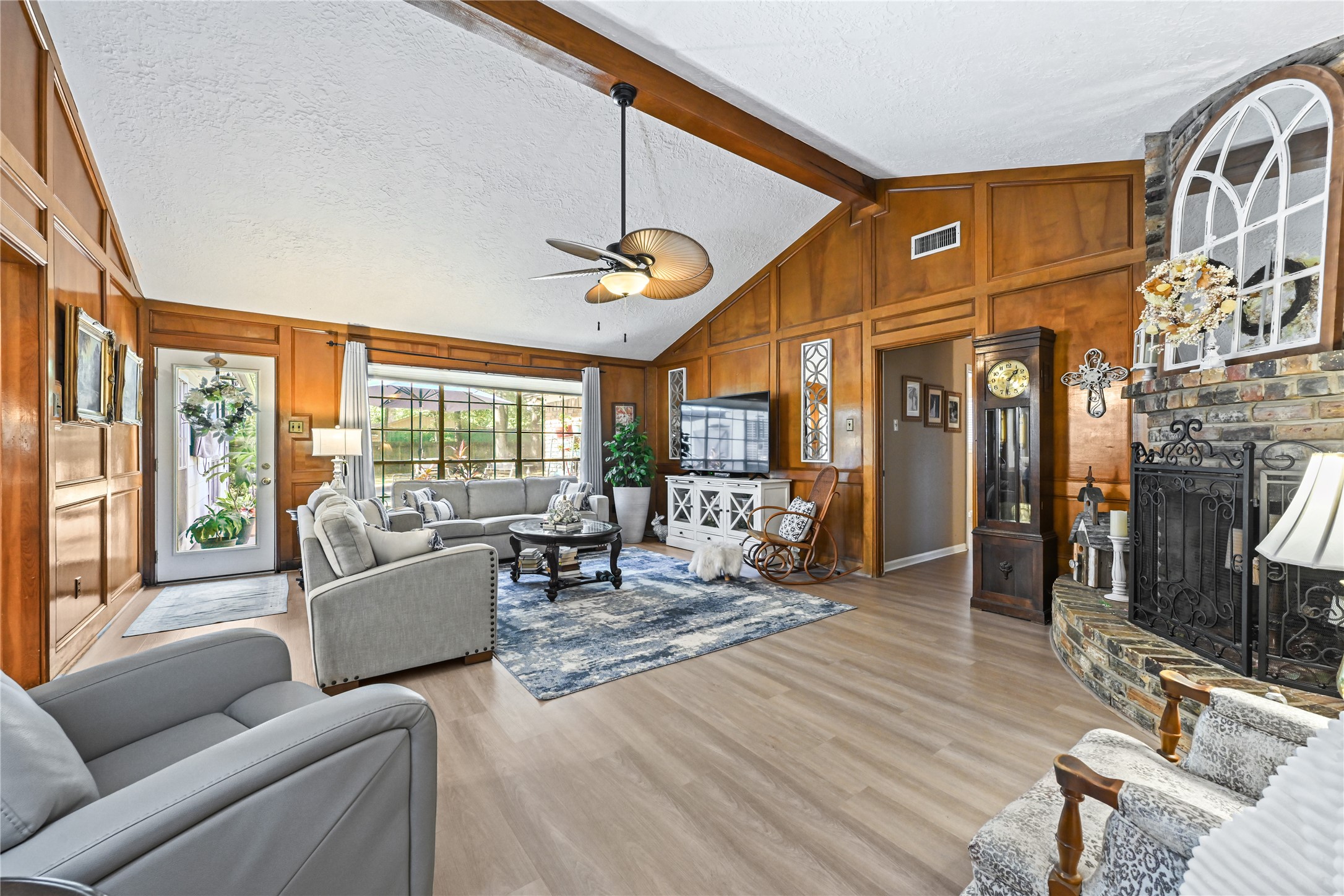 Spacious living room with vaulted ceilings, rich wood paneling, and a cozy brick fireplace. Features large windows for natural light, elegant furnishings, and a stylish ceiling fan, creating a warm and inviting atmosphere.