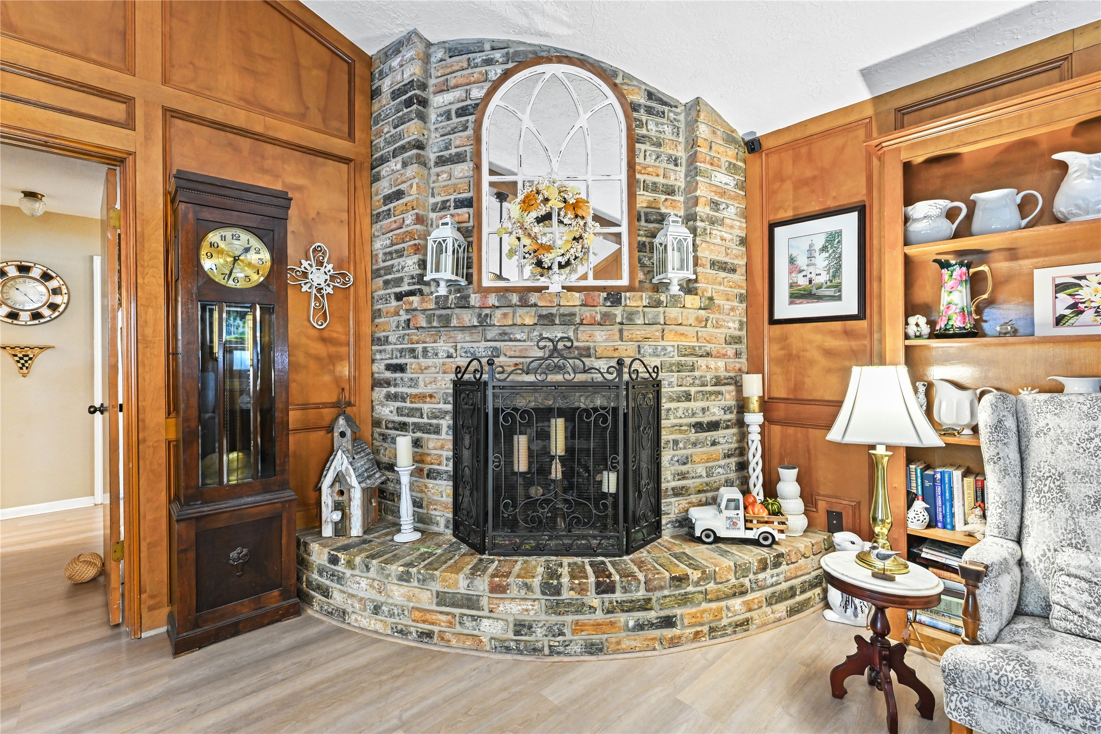 Charming living area featuring a rustic brick fireplace surrounded by rich wood paneling, a classic grandfather clock, and elegant built-in shelving.