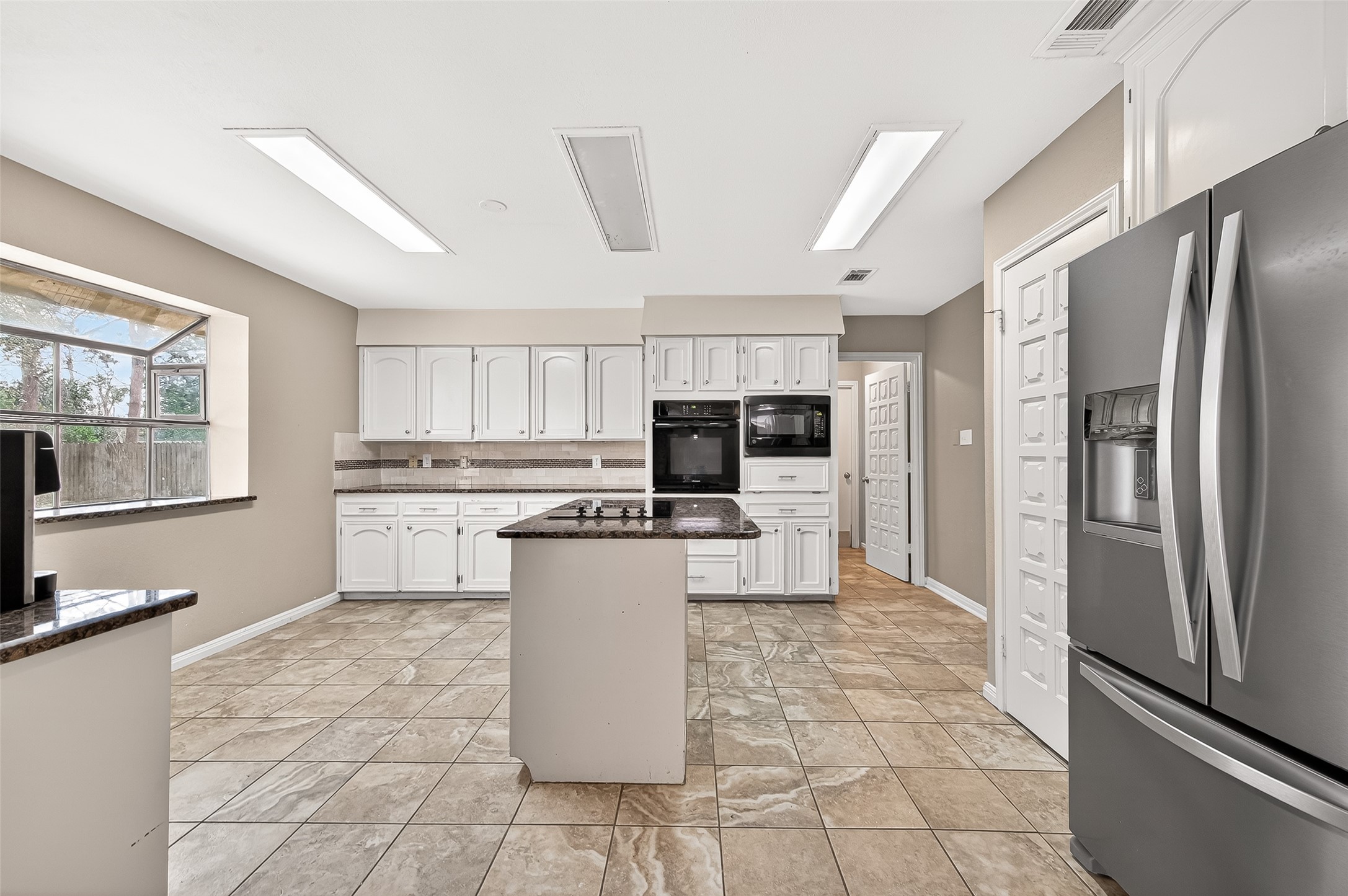 This large kitchen features modern appliances, ample white cabinetry, and a central island with a cooktop. Natural light flows through a large window, highlighting the tiled floor and a contemporary design perfect for cooking and entertaining.