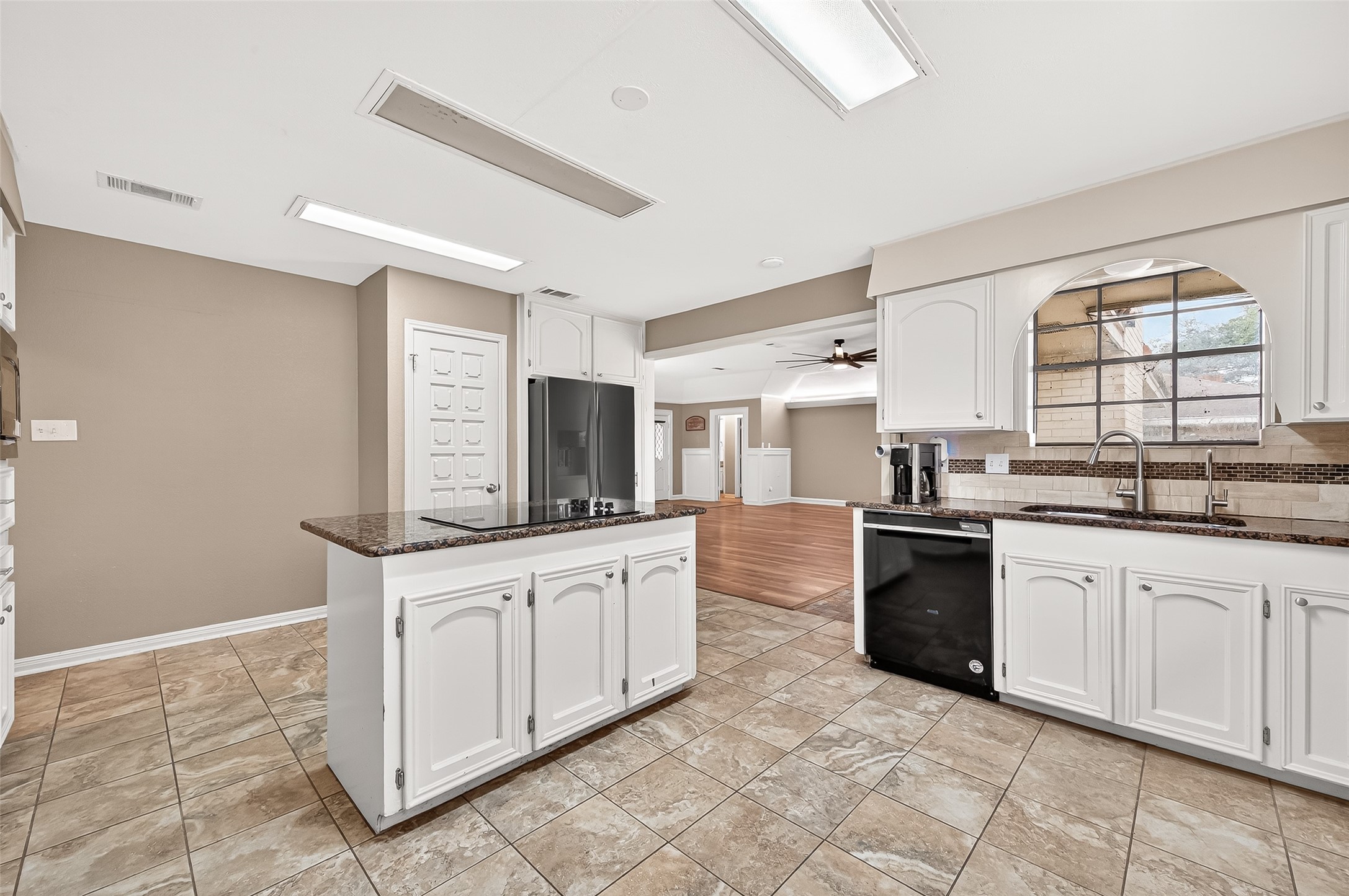 This photo showcases a modern kitchen with white cabinetry, granite countertops, and tile flooring. It features stainless steel appliances and an open layout leading to a living area with wood flooring and a ceiling fan. The space is bright and welcoming, ideal for entertaining.