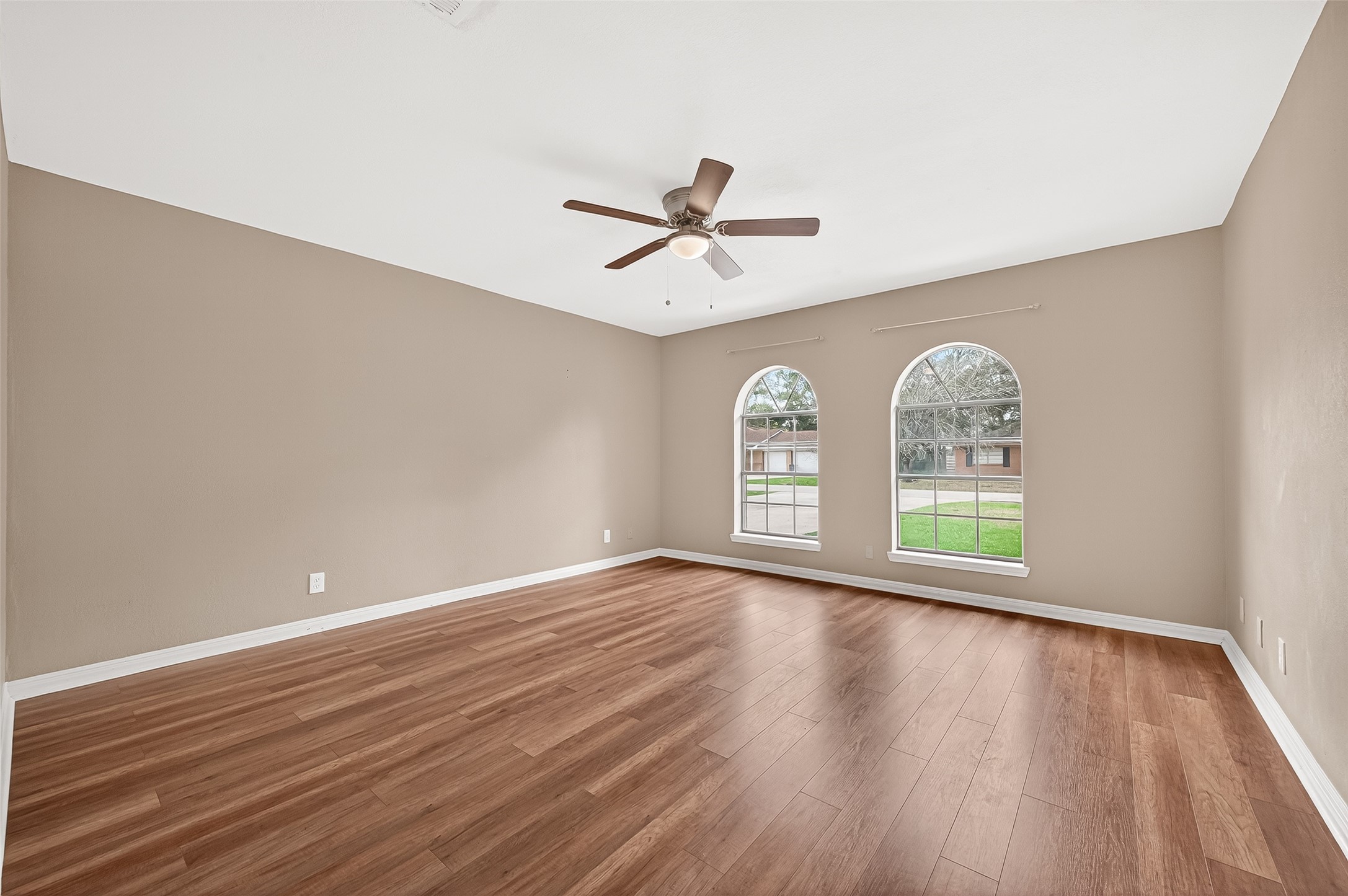 Primary bedroom with warm wood flooring, neutral walls, and a ceiling fan. Features two large arched windows providing plenty of natural light and a view of the front yard. Perfect for a cozy living area or bedroom.