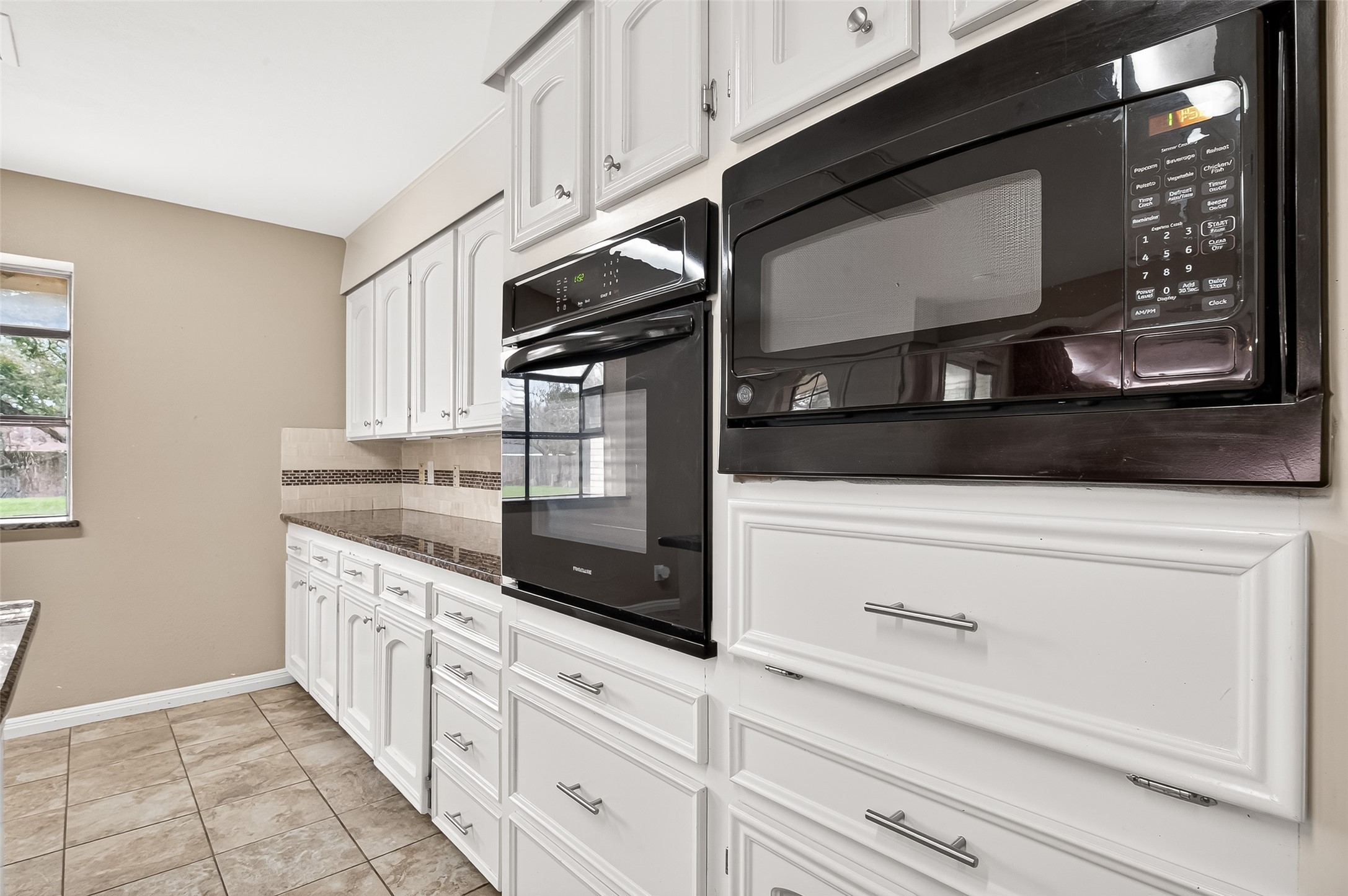 This kitchen features modern black appliances, including a built-in oven and microwave, set against crisp white cabinetry. The tile countertops and backsplash add a touch of elegance, while the large window provides ample natural light.