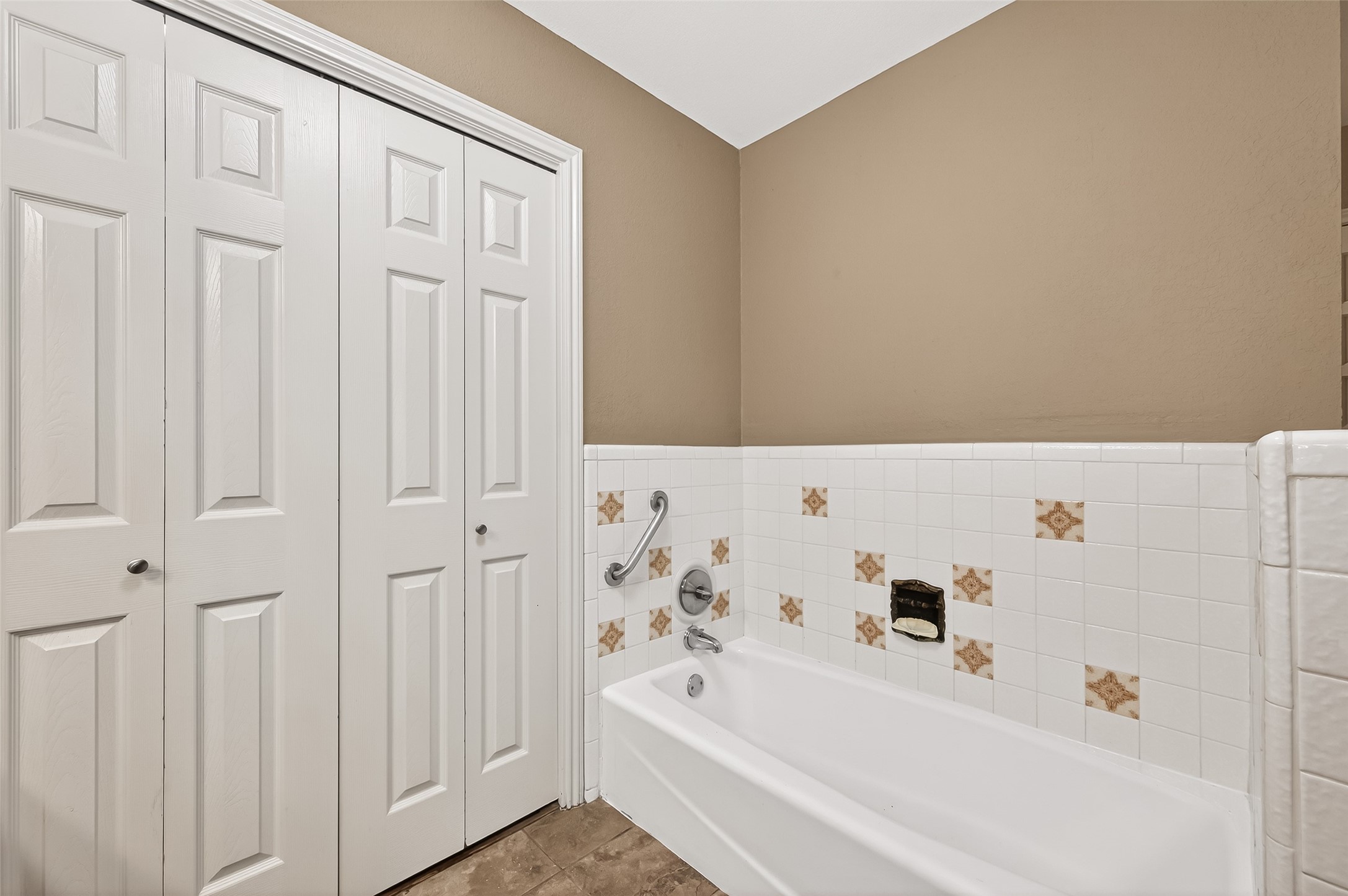 This bathroom features a bathtub with vintage tile accents, a grab bar for safety, and a closet with white paneled doors, offering both style and functionality.