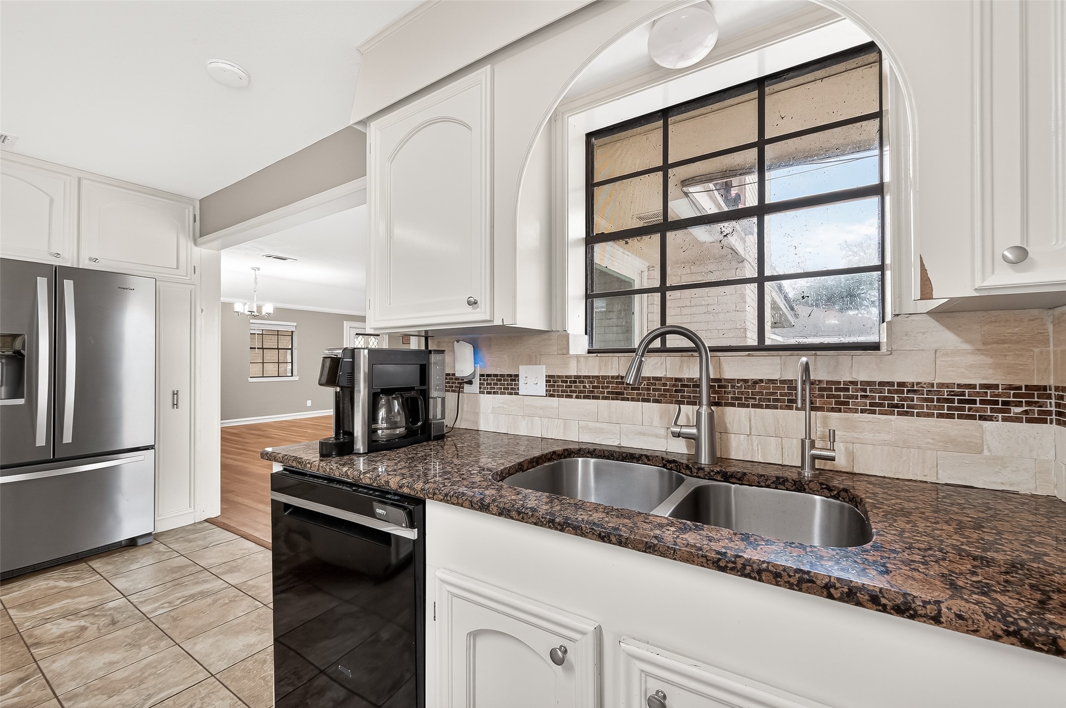Modern kitchen with granite countertops, dual sinks, and stainless steel appliances. Features a large window for natural light, white cabinetry, and a view into the adjacent dining area. Large kitchen window overlooking the backyard.