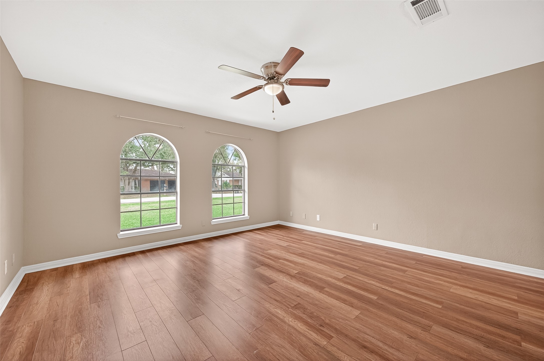 This spacious room features elegant wood flooring, two large arched windows providing ample natural light, neutral walls, and a ceiling fan for comfort.