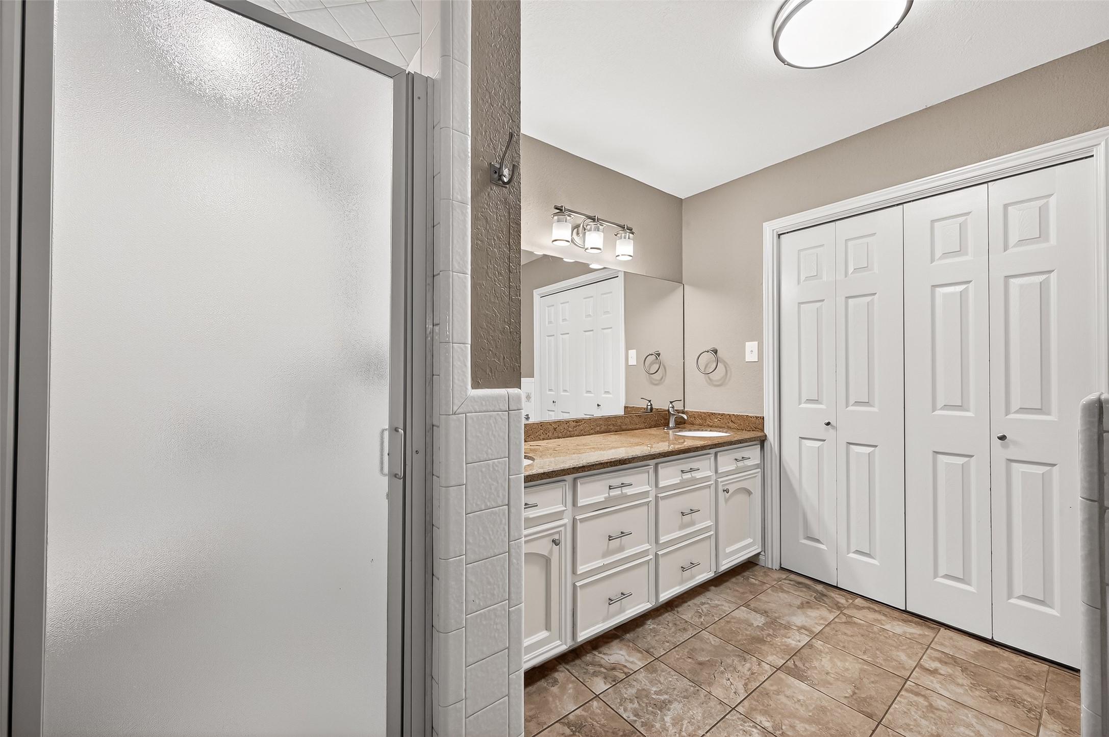 This bathroom features a modern frosted glass shower, double vanity with granite countertops, ample cabinet storage, and large closet doors, complemented by tile flooring and sleek lighting.