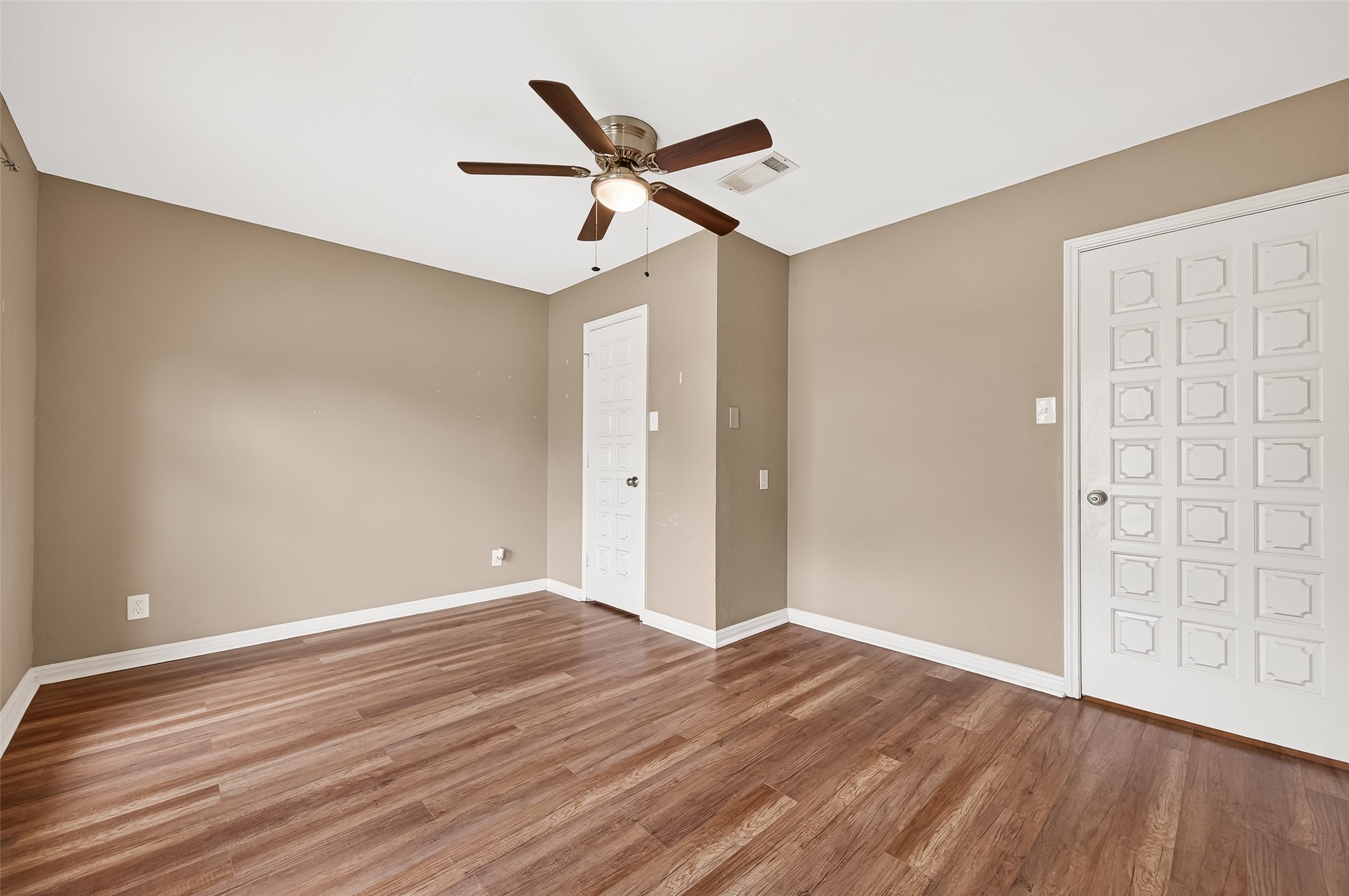 This room features warm beige walls, hardwood floors, and a ceiling fan. It's bright and inviting, with two doors for easy access. Ideal for a bedroom or home office.