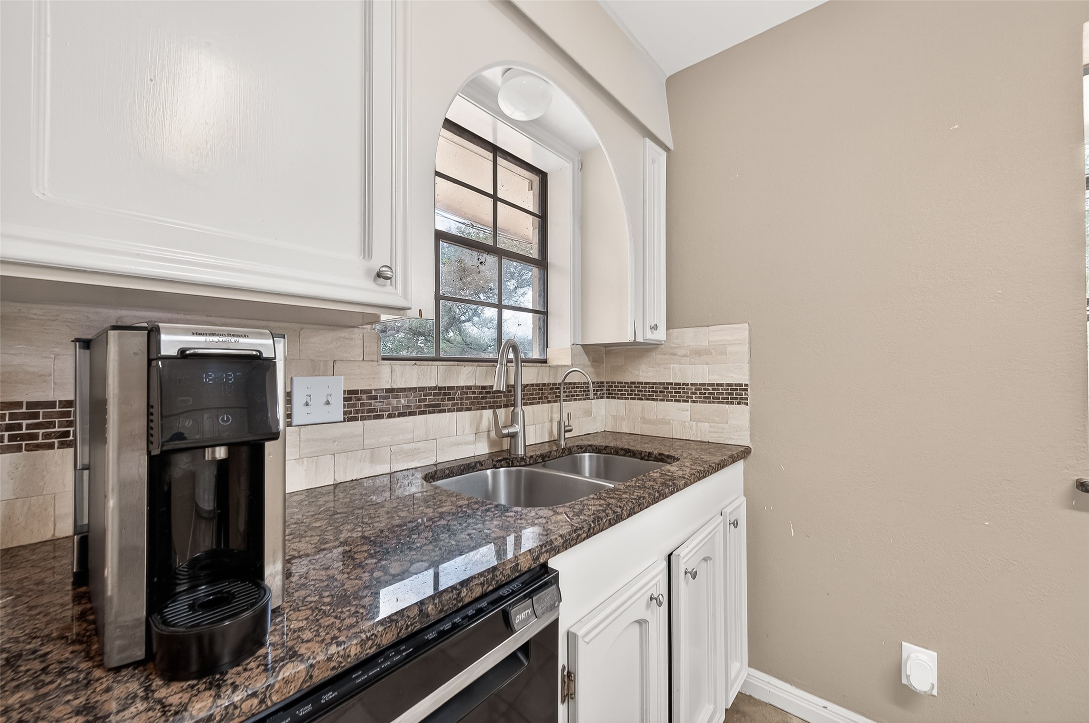 This kitchen features a sleek design with granite countertops, and a double sink. It includes white cabinetry, a tile backsplash, and a large window for natural light.