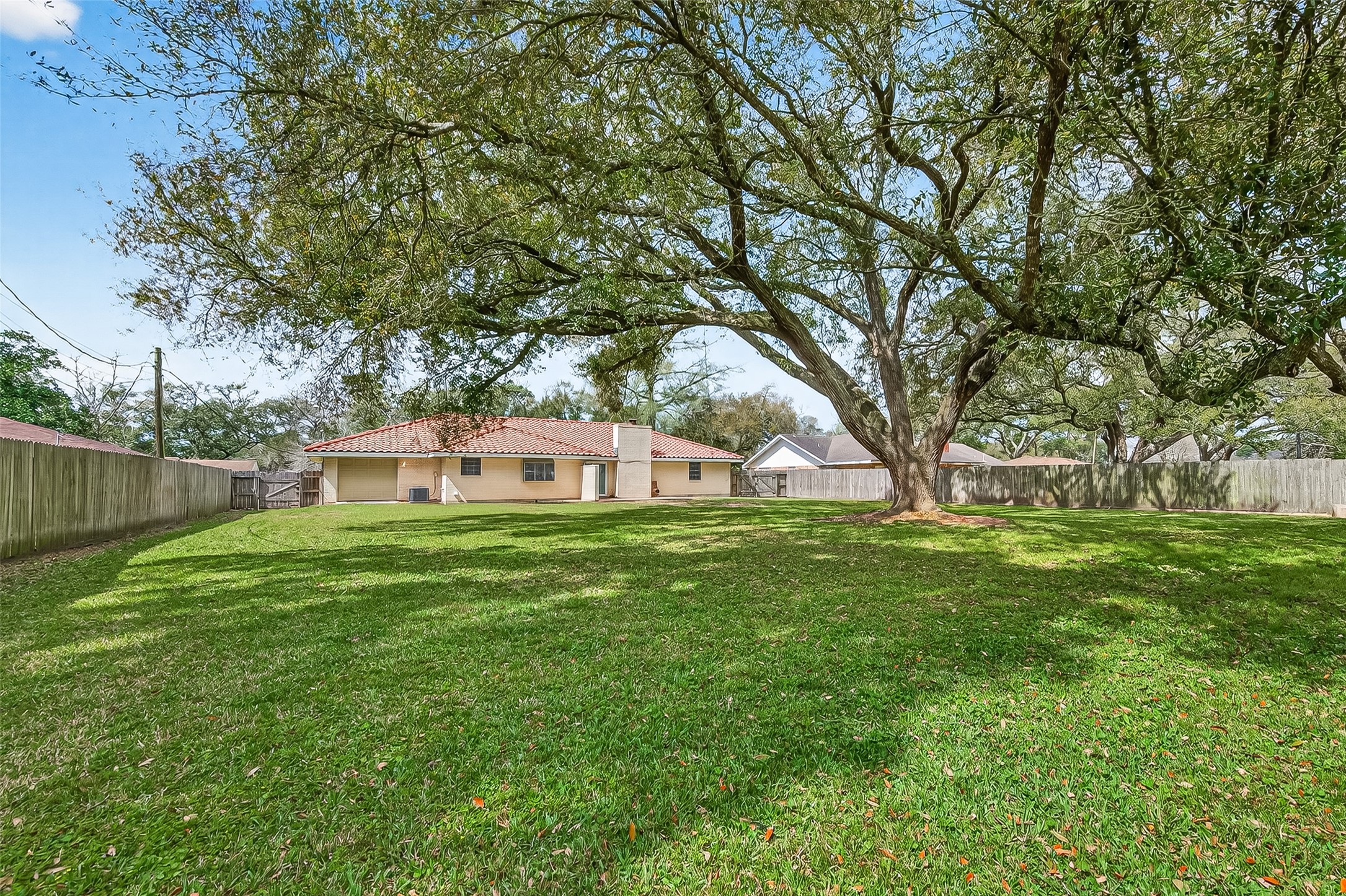 This photo showcases a spacious backyard with a large, mature tree providing ample shade. The area is fully fenced, offering privacy, and the view includes a single-story house with a red-tiled roof, making it ideal for outdoor activities and relaxation.
