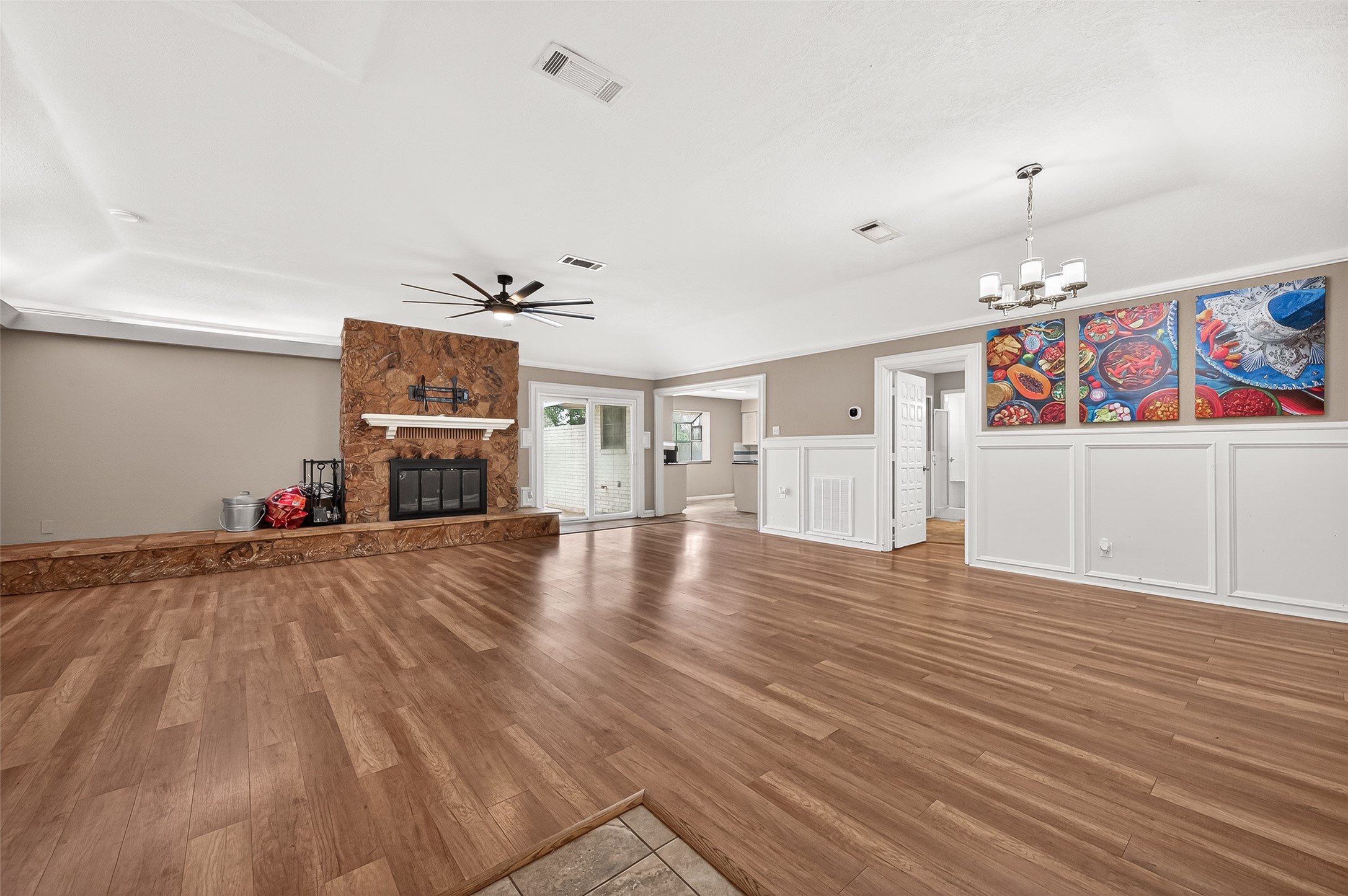 This spacious living area features hardwood flooring and a stone fireplace. It includes a ceiling fan, dining space with a modern chandelier, and vibrant wall art. The room opens to a bright kitchen, offering a welcoming, open-concept feel.