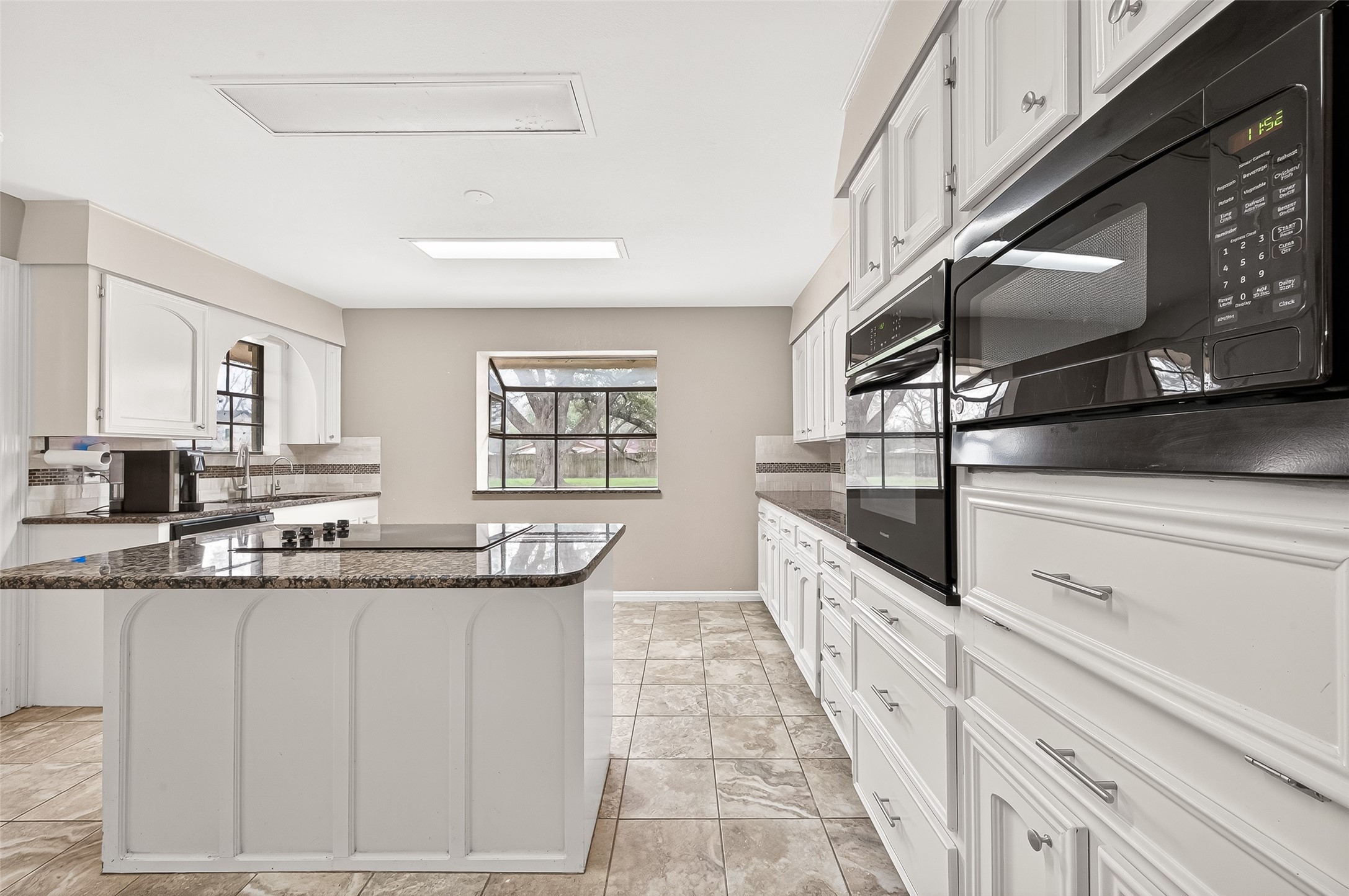 Spacious kitchen with a large island, granite countertops, and ample white cabinetry. Features modern appliances, tiled flooring, and a window with a pleasant view.