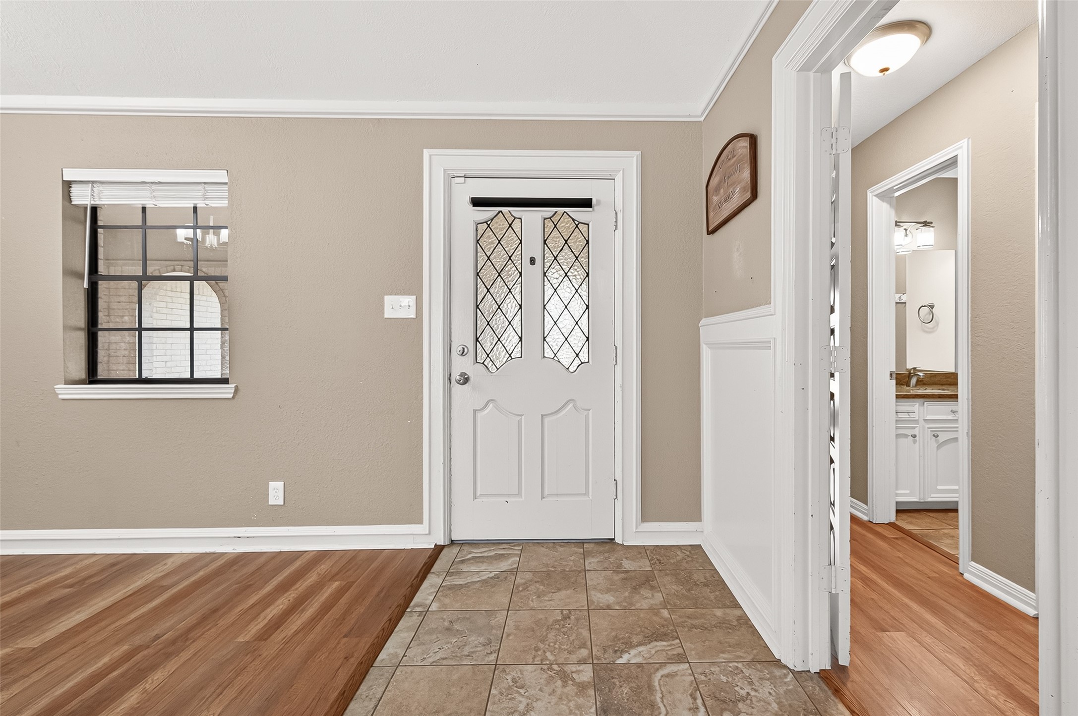 This photo showcases a cozy entryway with a white door featuring decorative glass panels. The space has a mix of tile and wood flooring, beige walls, and wainscoting.