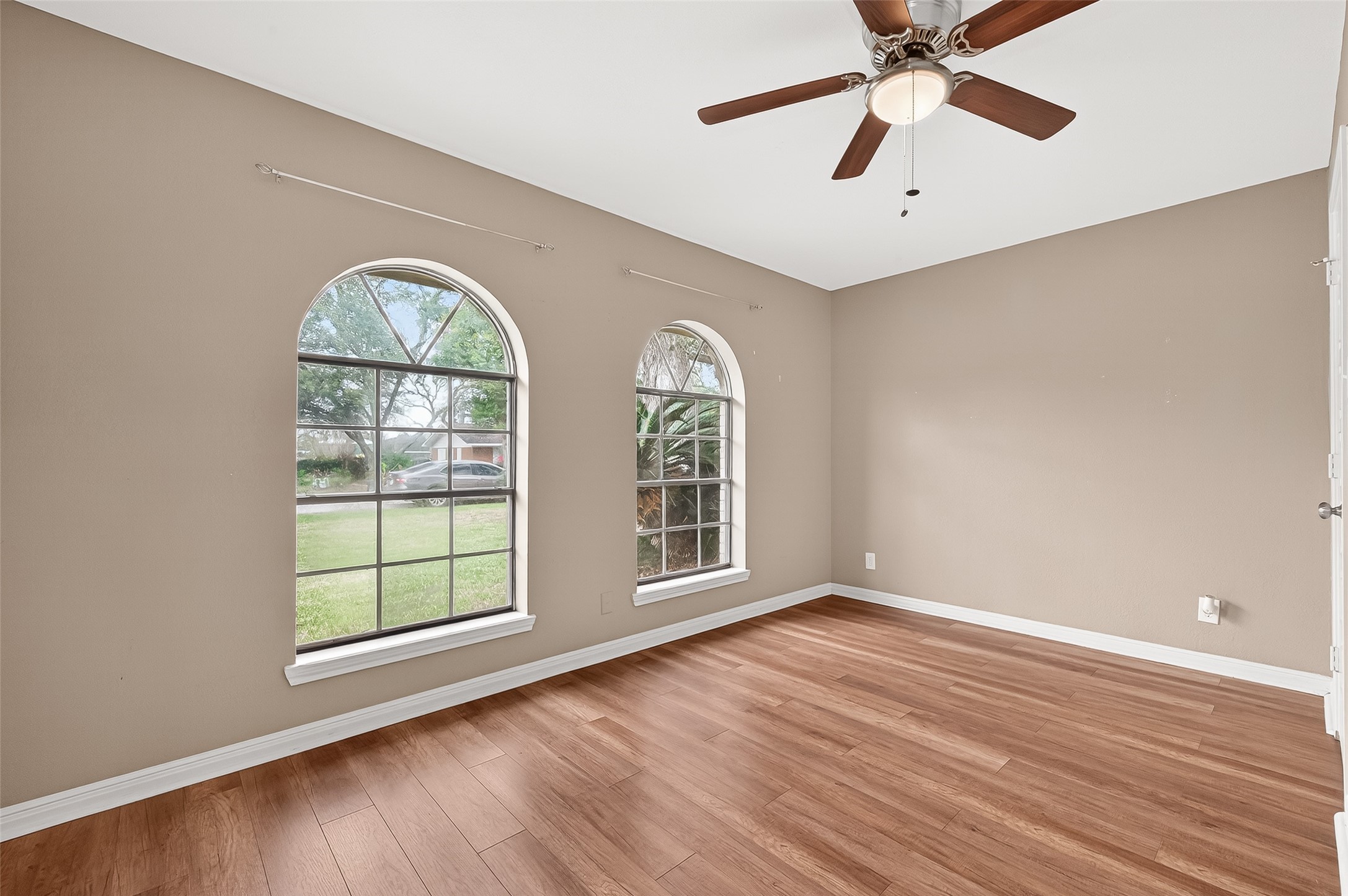 This room features two large arched windows, allowing natural light to fill the space. It has warm wood flooring, neutral walls, and a ceiling fan, creating a cozy and inviting atmosphere.