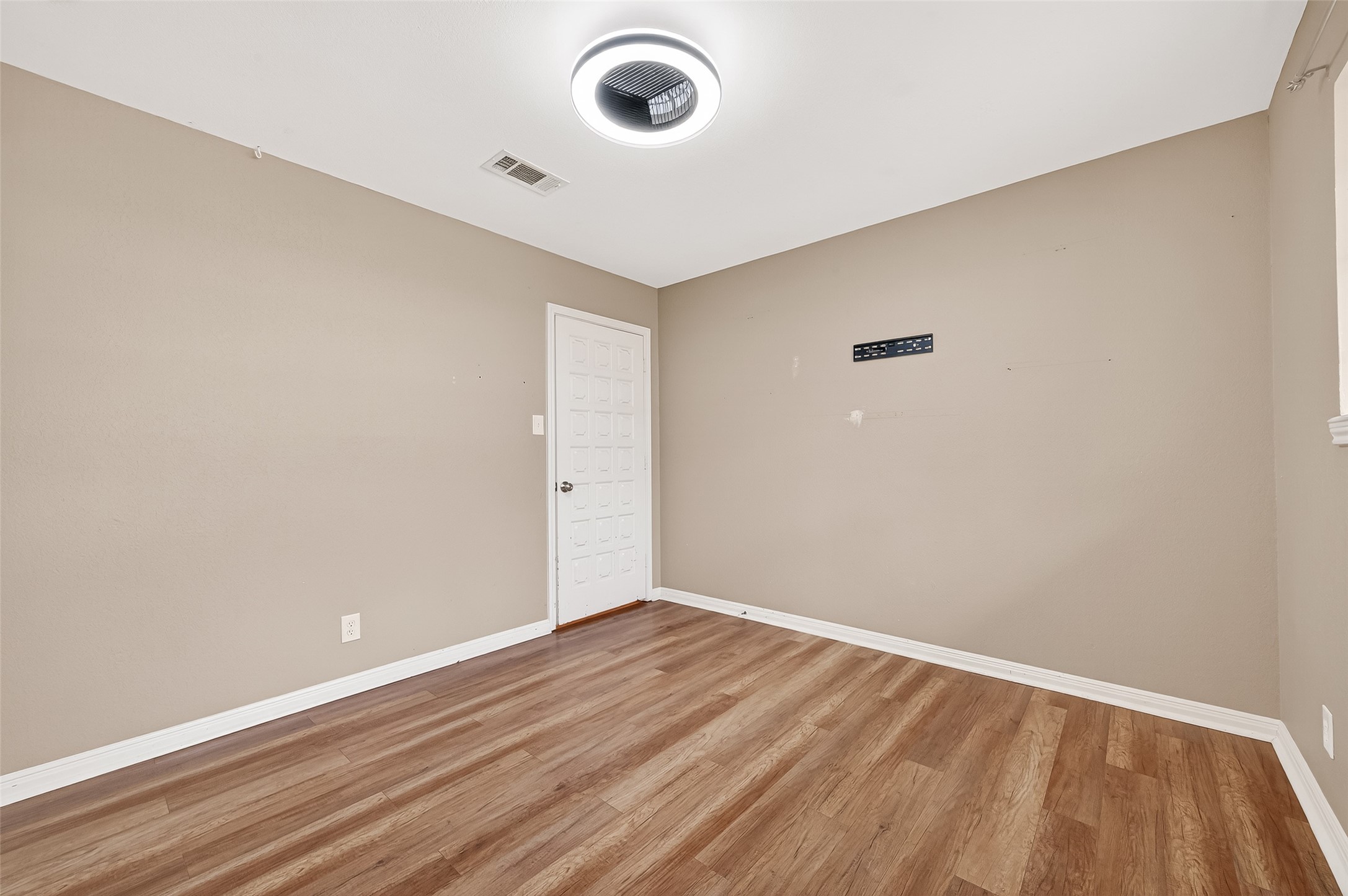 This photo shows a simple, unfurnished room with beige walls and wood flooring. It features a modern ceiling light and a door, creating a clean and versatile space ready for customization.