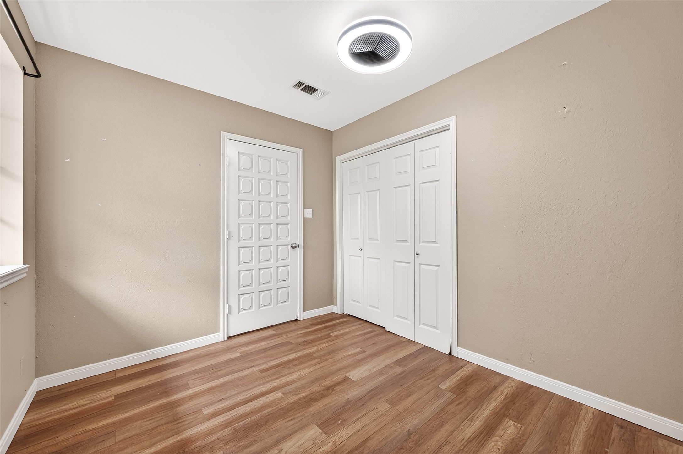 This room features light brown walls and laminate wood flooring, with a window allowing natural light. It includes a modern ceiling light and two white doors, one with a geometric design and the other a closet, providing ample storage space.