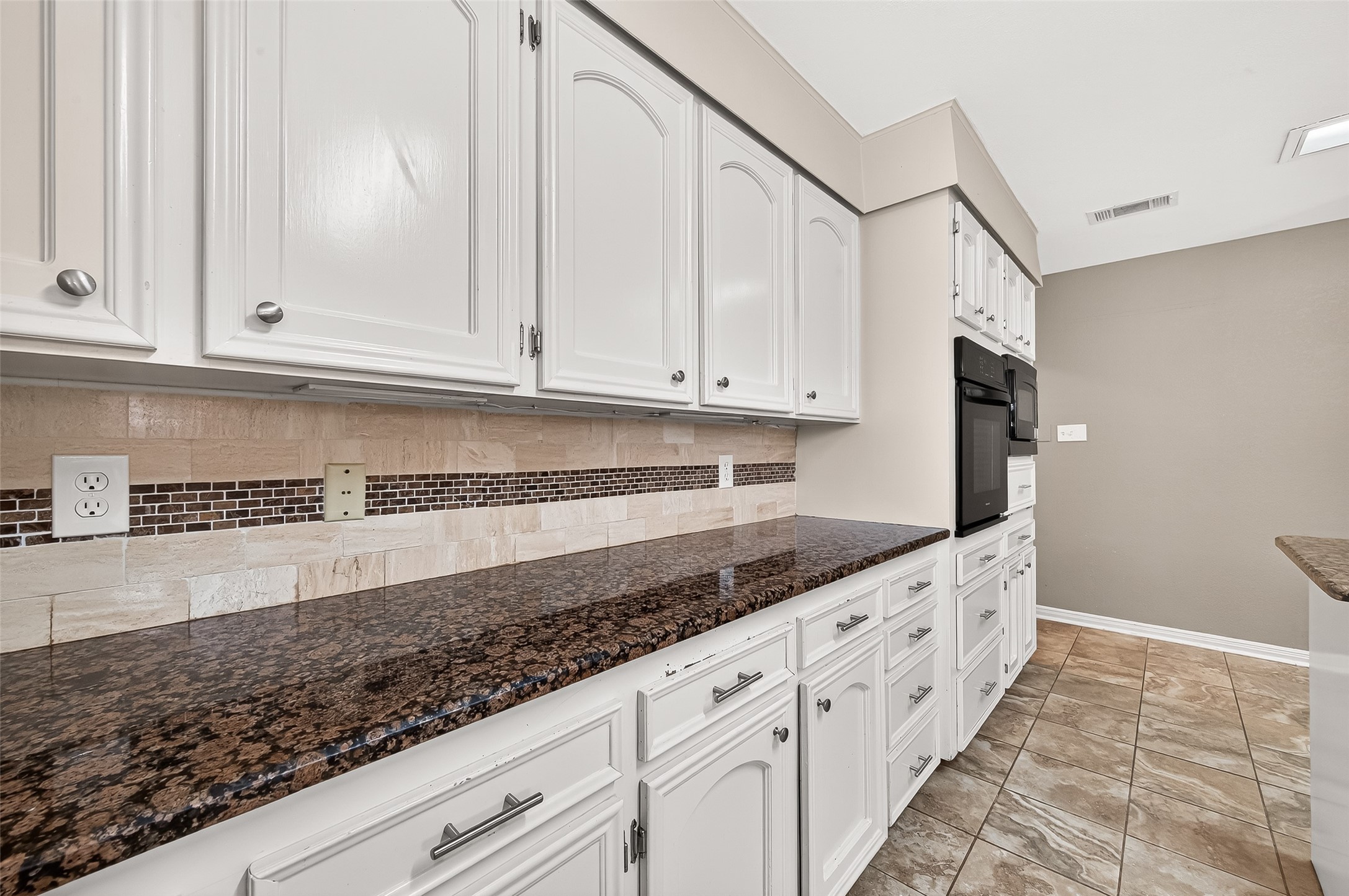 This kitchen features elegant white cabinetry with ample storage, stunning dark granite countertops, and a stylish tile backsplash. The space is complemented by tiled flooring and built-in appliances, offering a modern and functional layout.