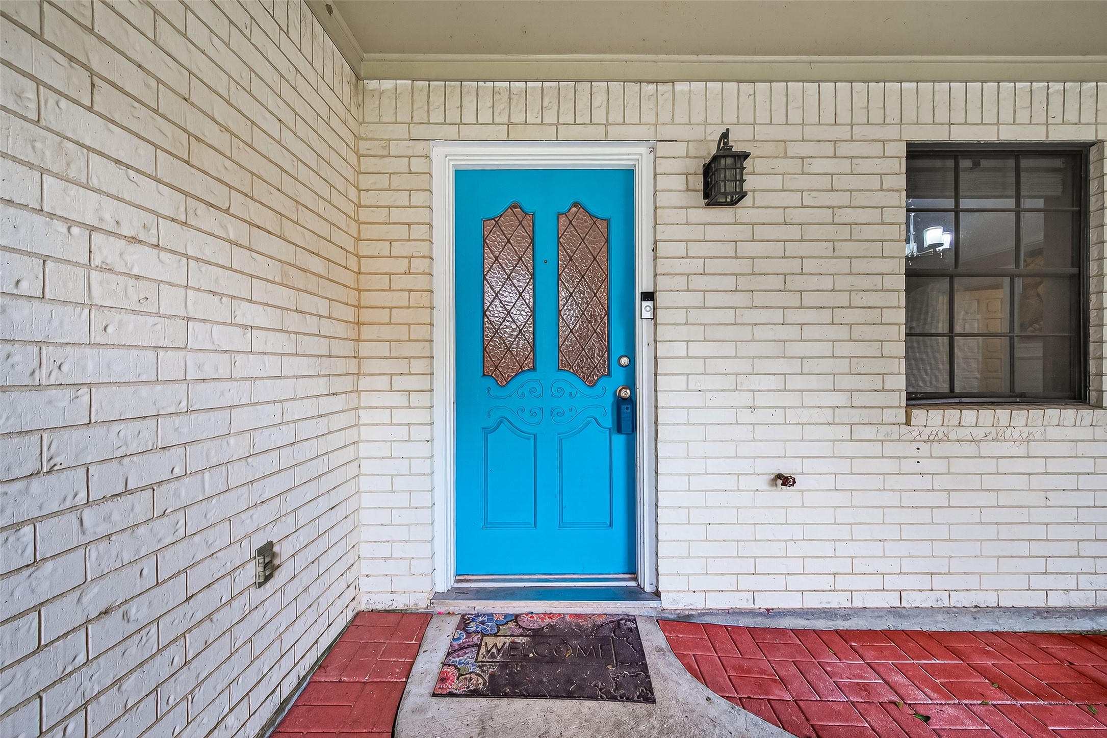 This photo showcases a welcoming front entrance with a vibrant blue door set in a white brick wall. The entrance features a decorative glass panel, a black outdoor light fixture, and a red brick pathway leading to a "Welcome" mat. A window with black trim is visible beside the door.