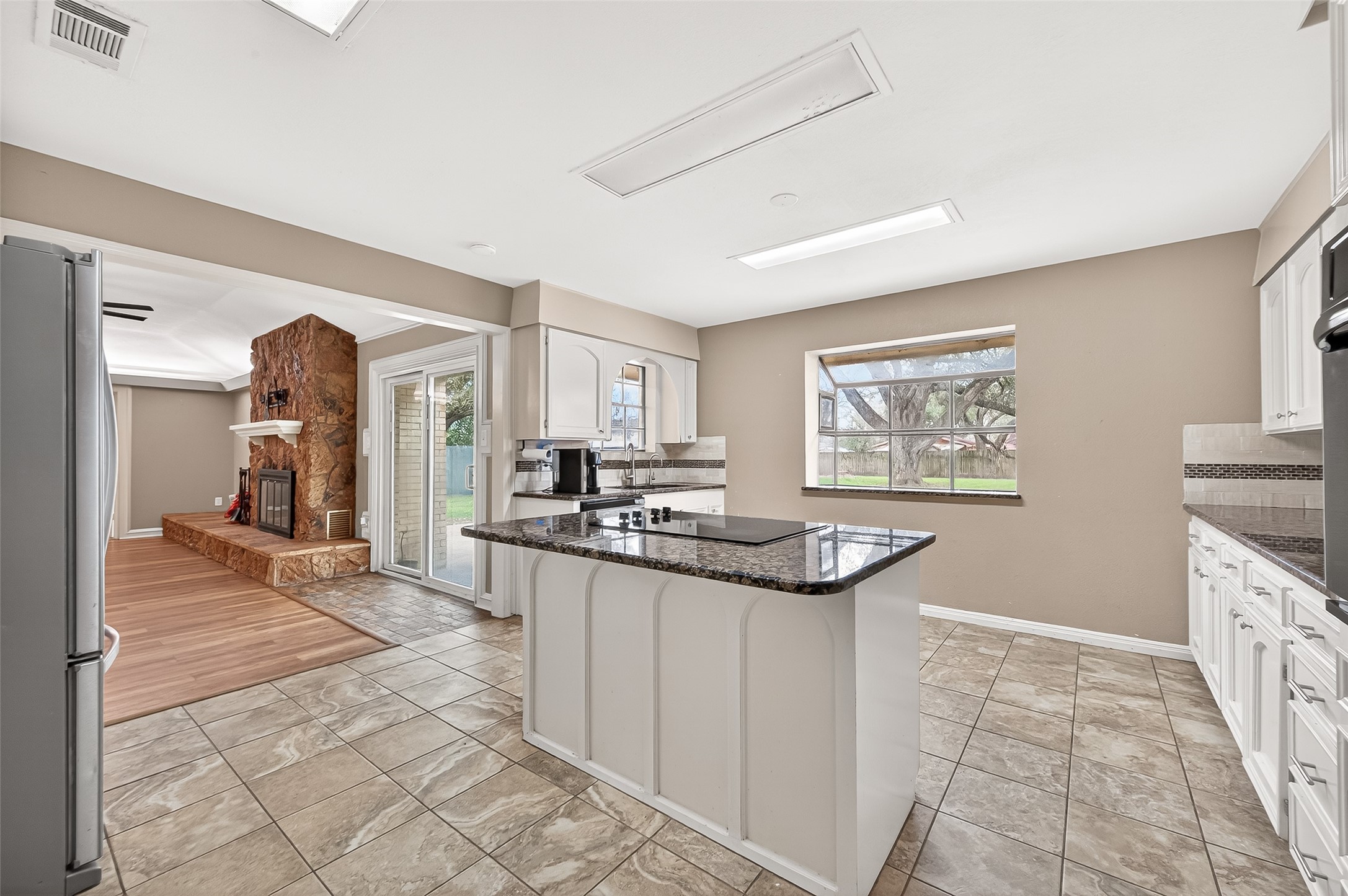 This kitchen features a central island with a cooktop, granite countertops, and ample cabinetry. It opens to a cozy living area with a stone fireplace and sliding doors leading to the backyard, offering natural light and a seamless flow for entertaining.