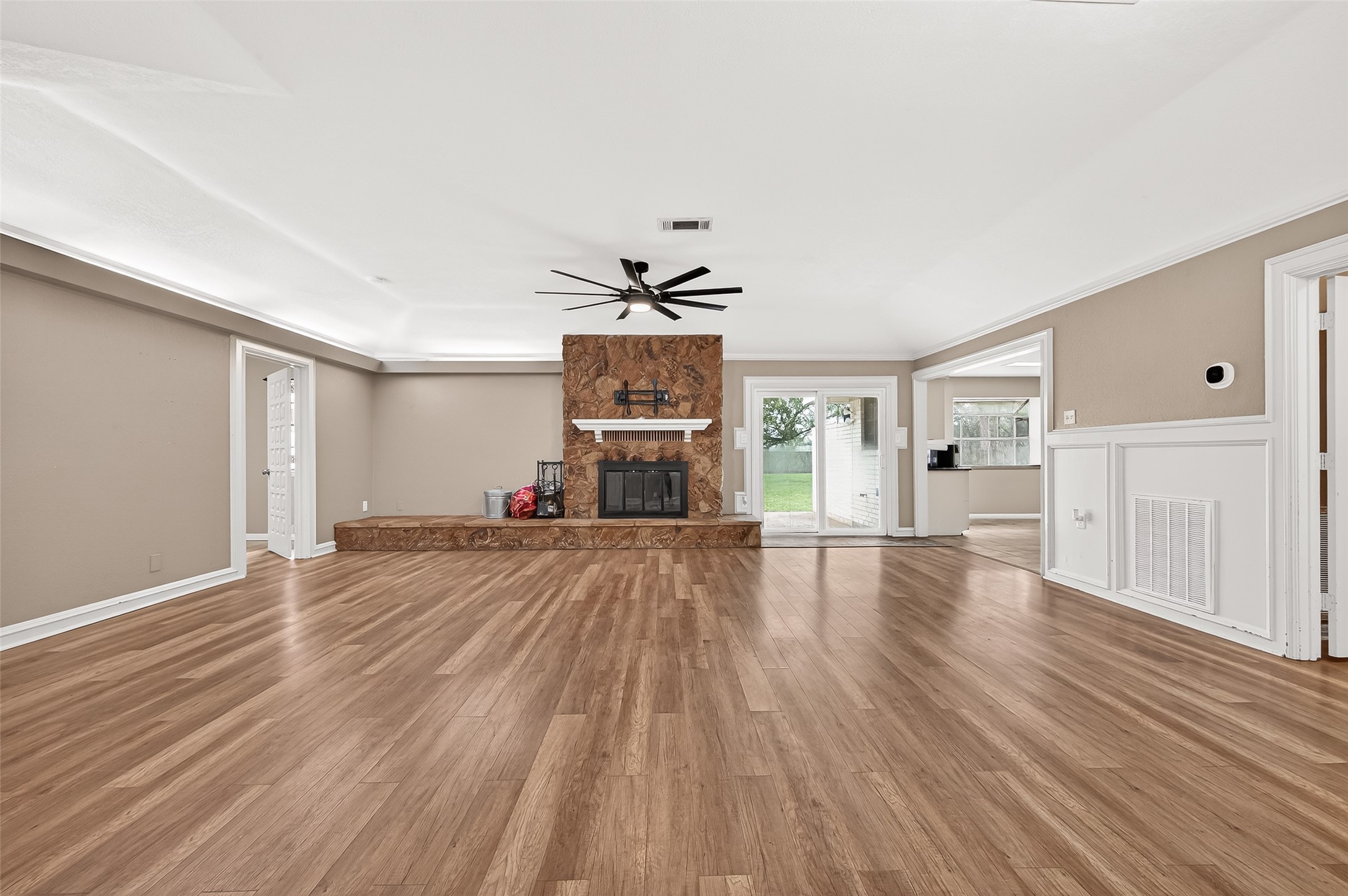 Spacious living room with hardwood floors, a central stone  wood burning fireplace, and large windows offering natural light. A ceiling fan adds comfort, while sliding doors lead to a lush backyard, perfect for indoor-outdoor living.