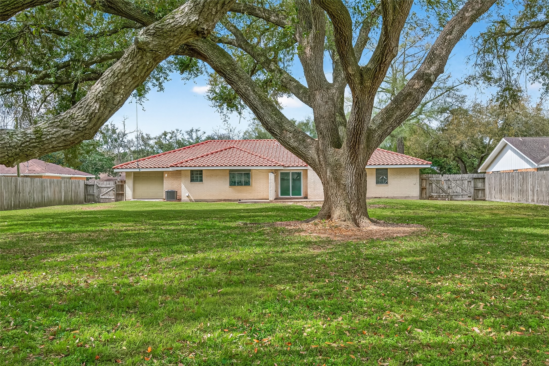 This photo features a charming single-story home with a red-tiled roof, set in a spacious, well-maintained backyard. A large, mature tree provides shade, and the yard is enclosed by a wooden fence, offering privacy and ample space for outdoor activities.