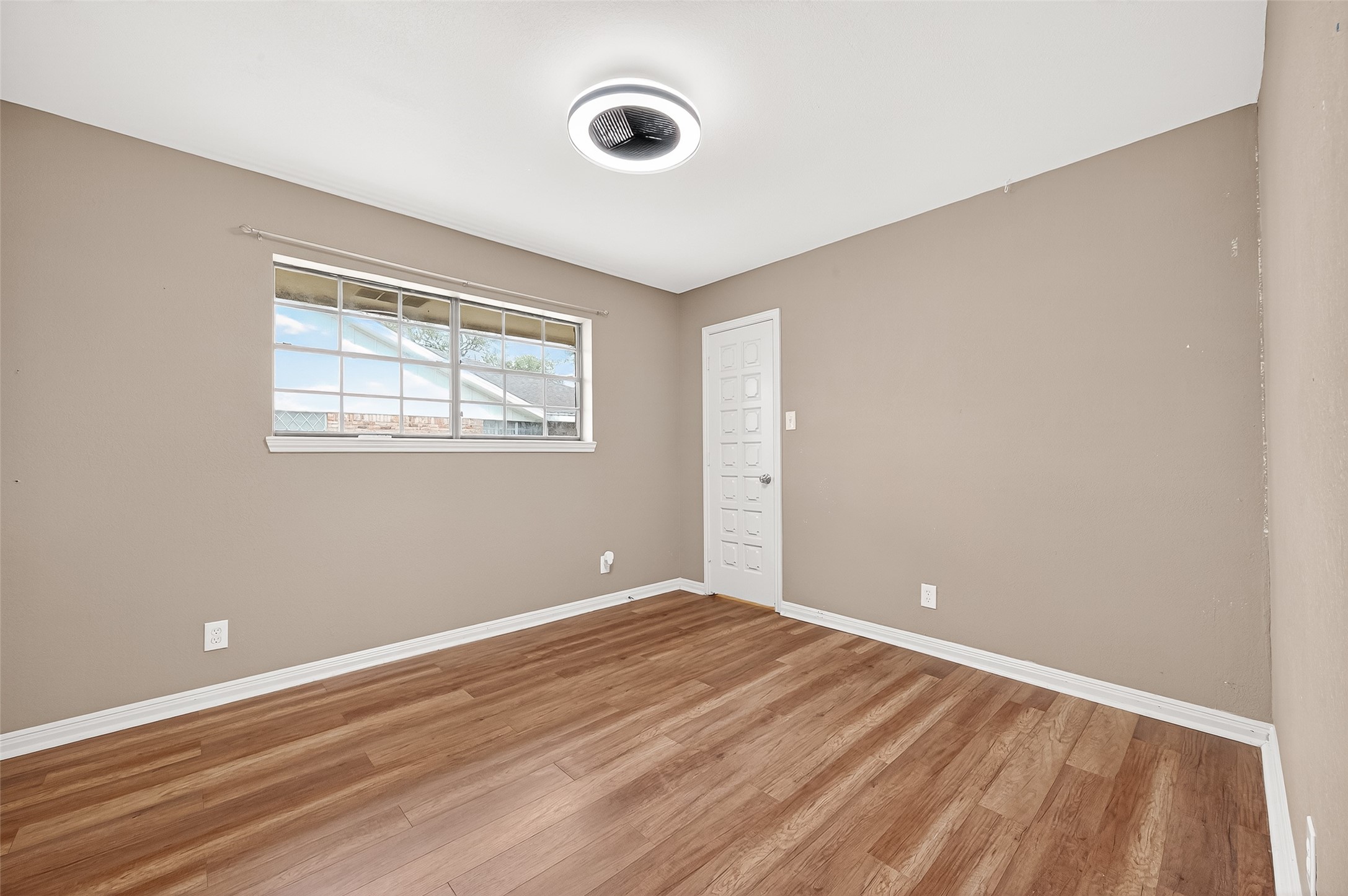 This room features neutral-colored walls, wood flooring, and a large window for natural light. The modern ceiling light and simple door add to its clean, fresh look.