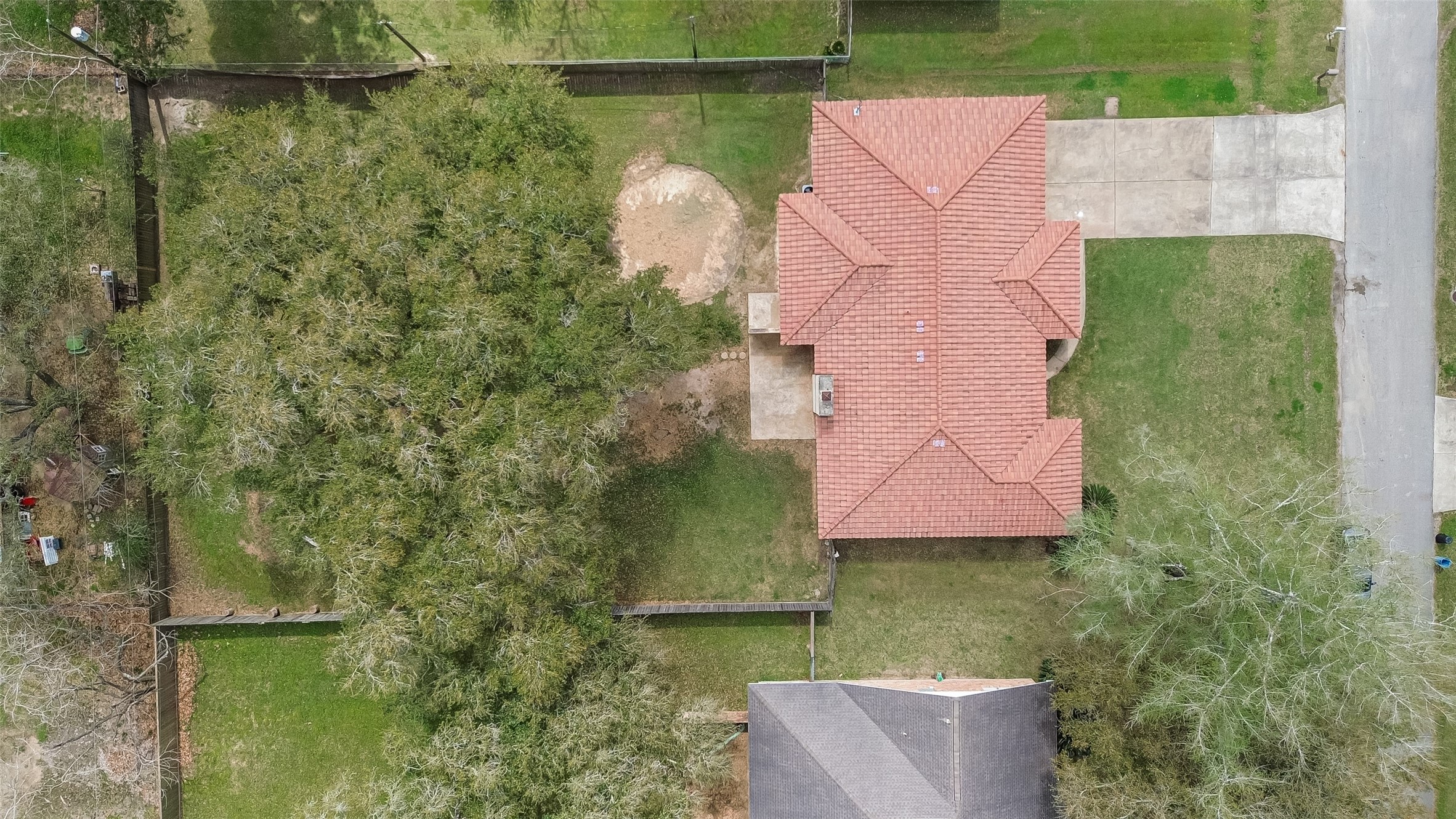 This aerial view shows a single-story home with a red-tiled roof and a spacious driveway. The property features a large fenced backyard with mature trees, providing ample shade and privacy. The house is situated in a residential area with nearby green spaces.
