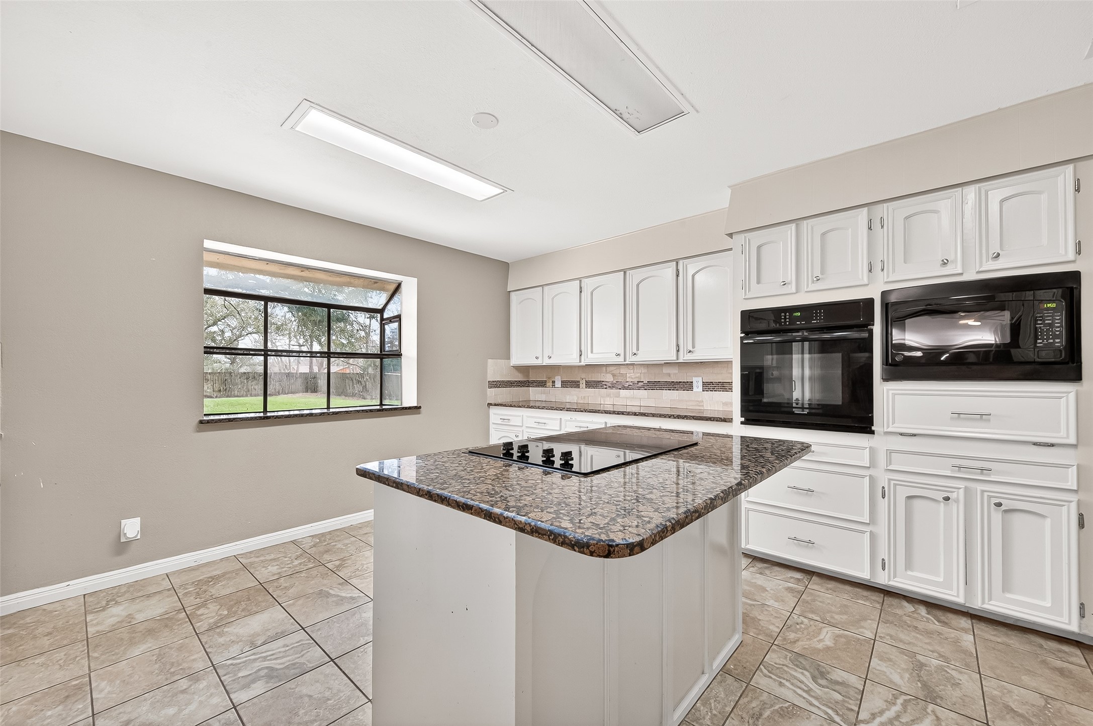 This spacious kitchen features a large window providing ample natural light, granite countertops, white cabinetry, and modern appliances, including a built-in oven and microwave. The central island offers additional prep space and a cooktop, perfect for culinary enthusiasts.