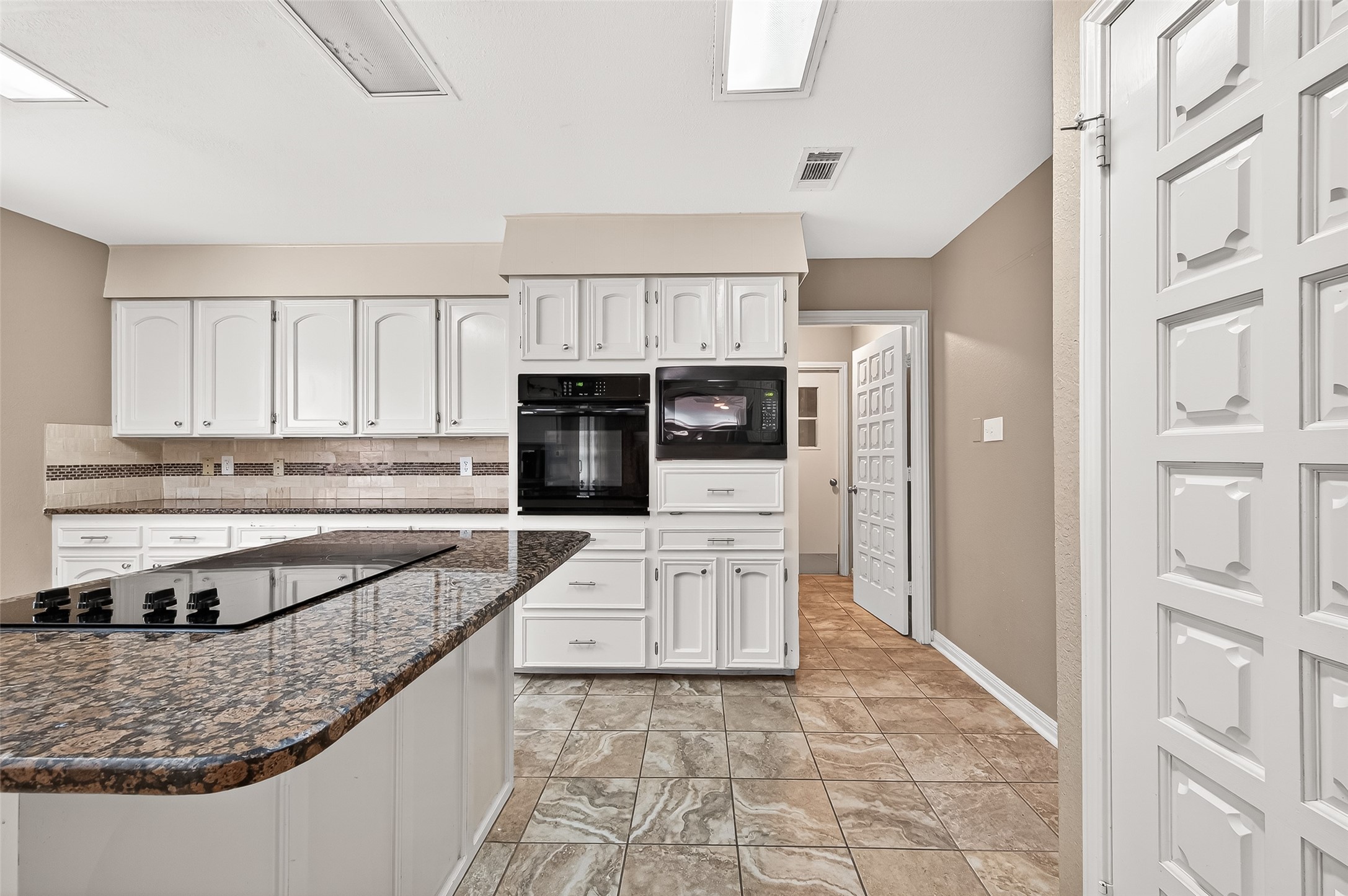 This kitchen features white cabinetry, a built-in oven and microwave, and a granite countertop island with a cooktop. The neutral tile flooring and backsplash add a classic touch, while the paneled doors provide a cohesive look.