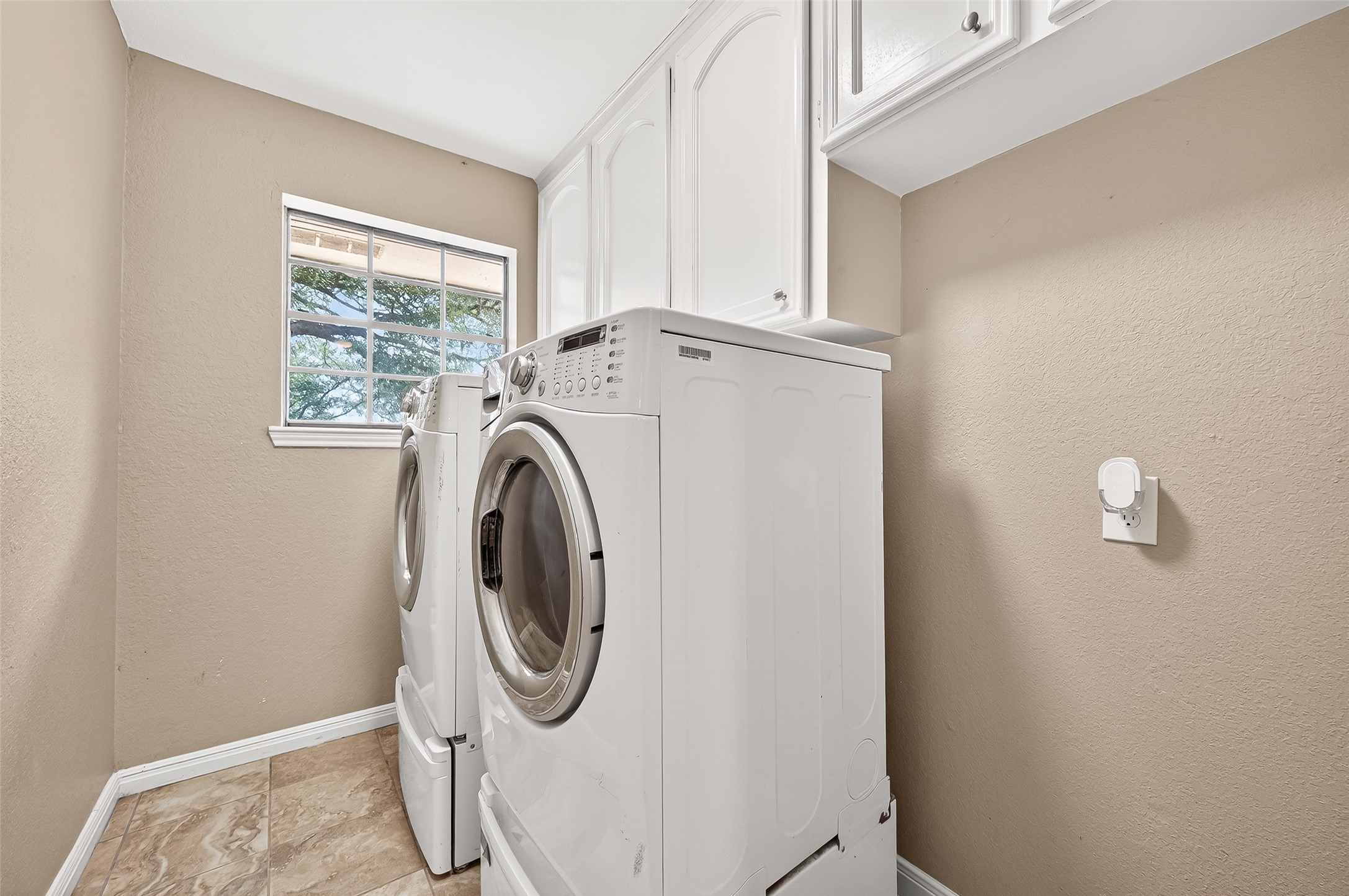 This photo shows a compact laundry room with a front-loading washer and dryer (washer and dryer not included). It features beige walls, white cabinetry for storage, and a window providing natural light. The floor is tiled, offering easy maintenance.