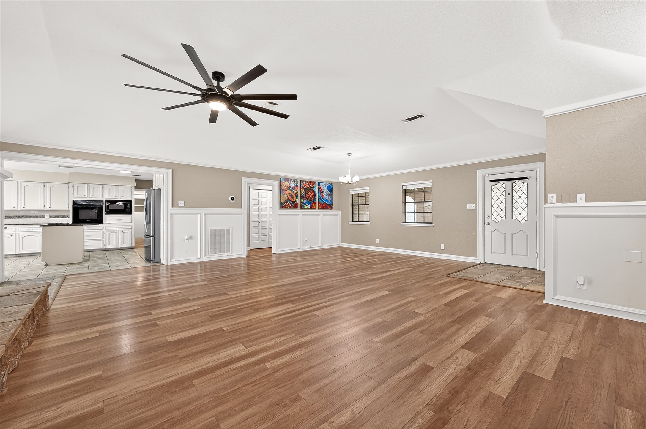 Living area with neutral walls. Features a modern ceiling fan and stylish wainscoting. The open layout leads to a bright kitchen with white cabinetry.