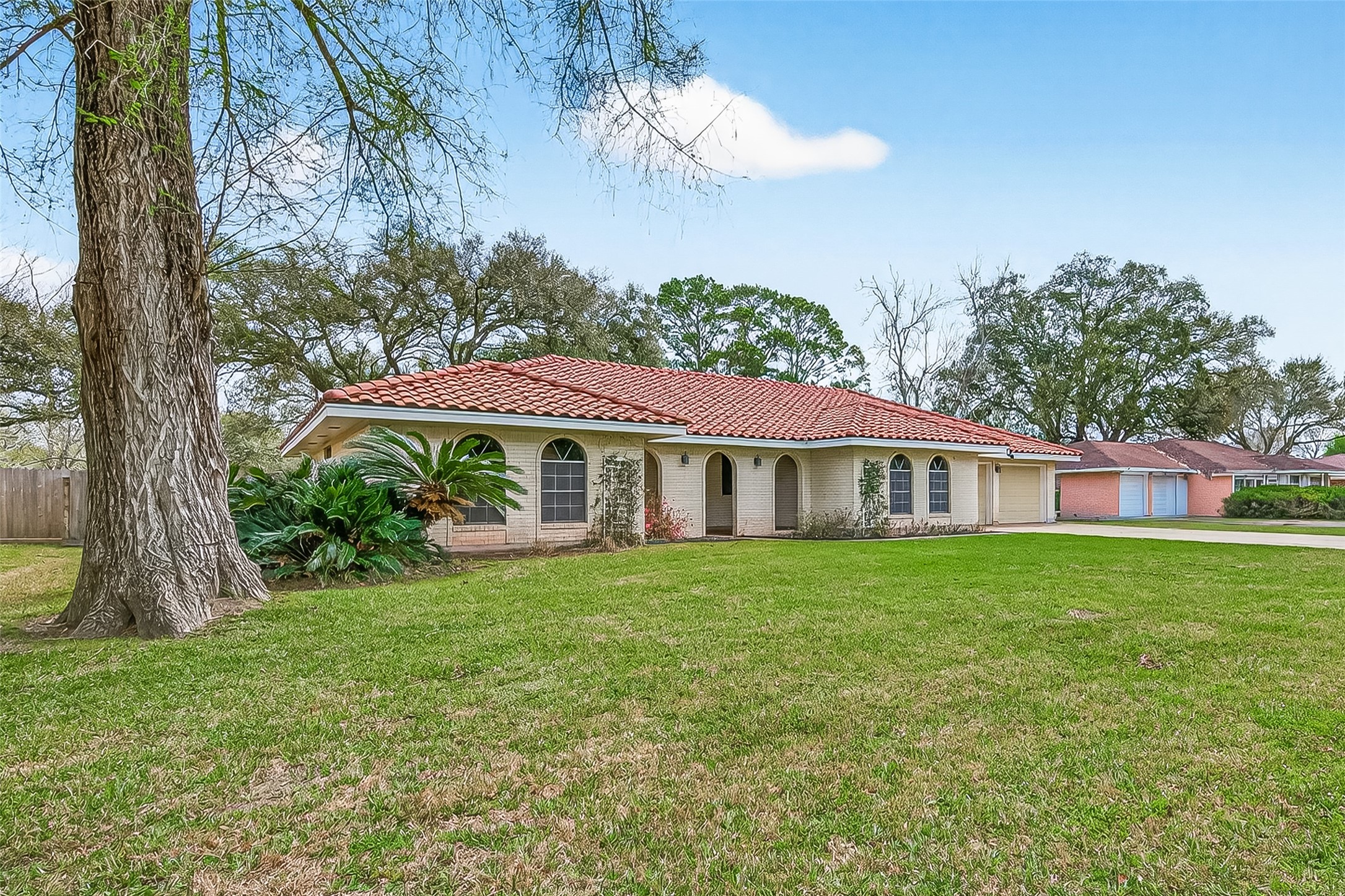 This charming home features a red-tiled roof and light brick exterior, set on a spacious, grassy lot with mature trees. The curved windows and arched entryway add character
