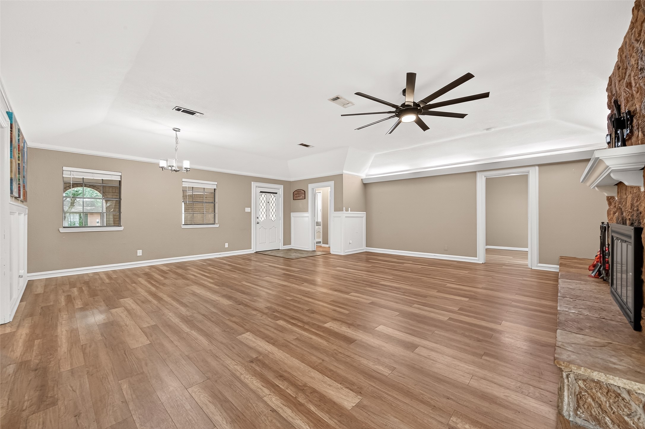 Spacious living room with wood flooring, neutral walls, and a ceiling fan. Features a stone fireplace and ample natural light from multiple windows. Includes an elegant chandelier and access to adjacent rooms.