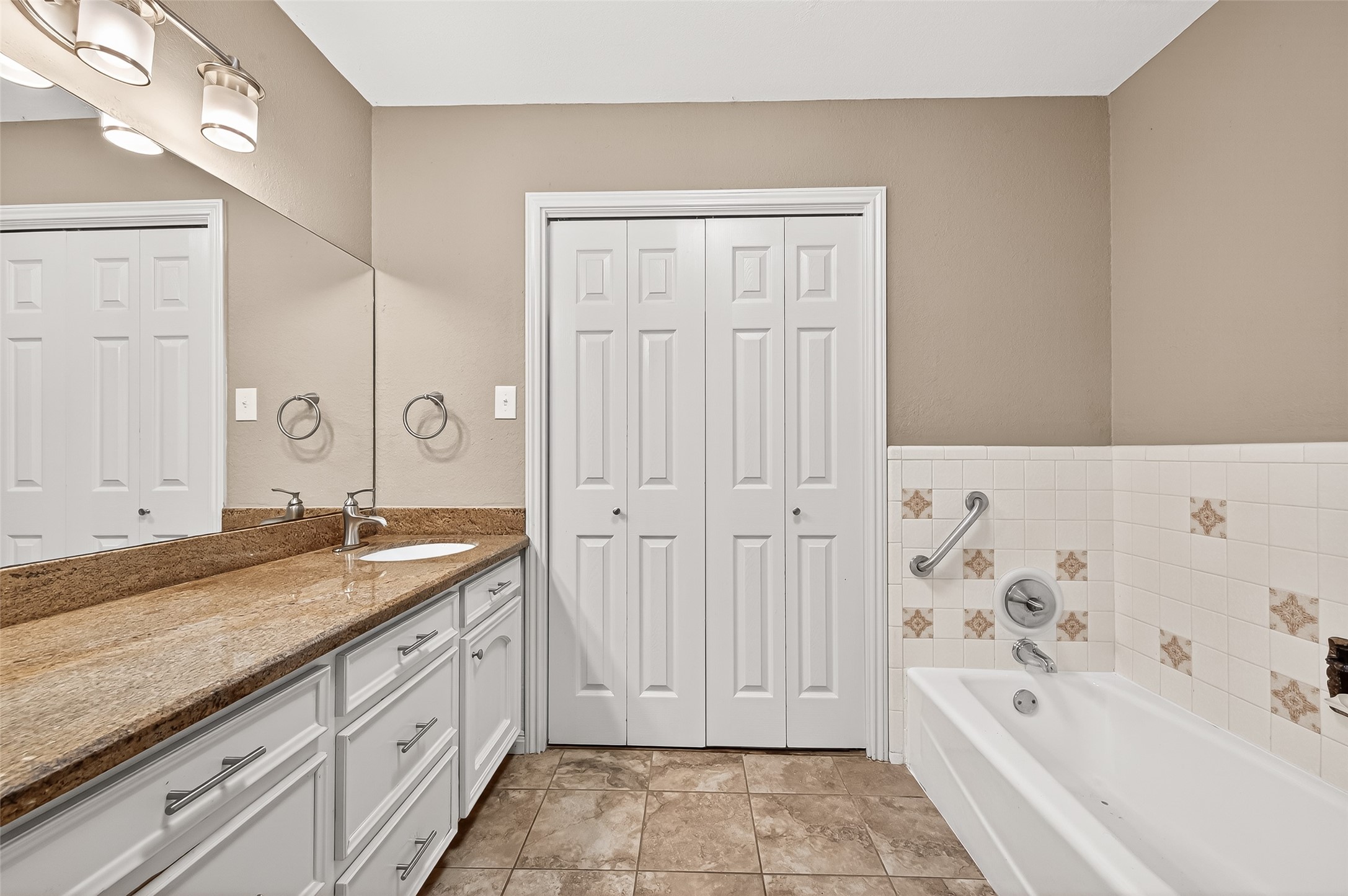 This bathroom features a long granite countertop with a single sink, ample storage with white cabinets, a tiled bathtub with a grab bar, and a double door closet. The decor is neutral, providing a clean and spacious feel.