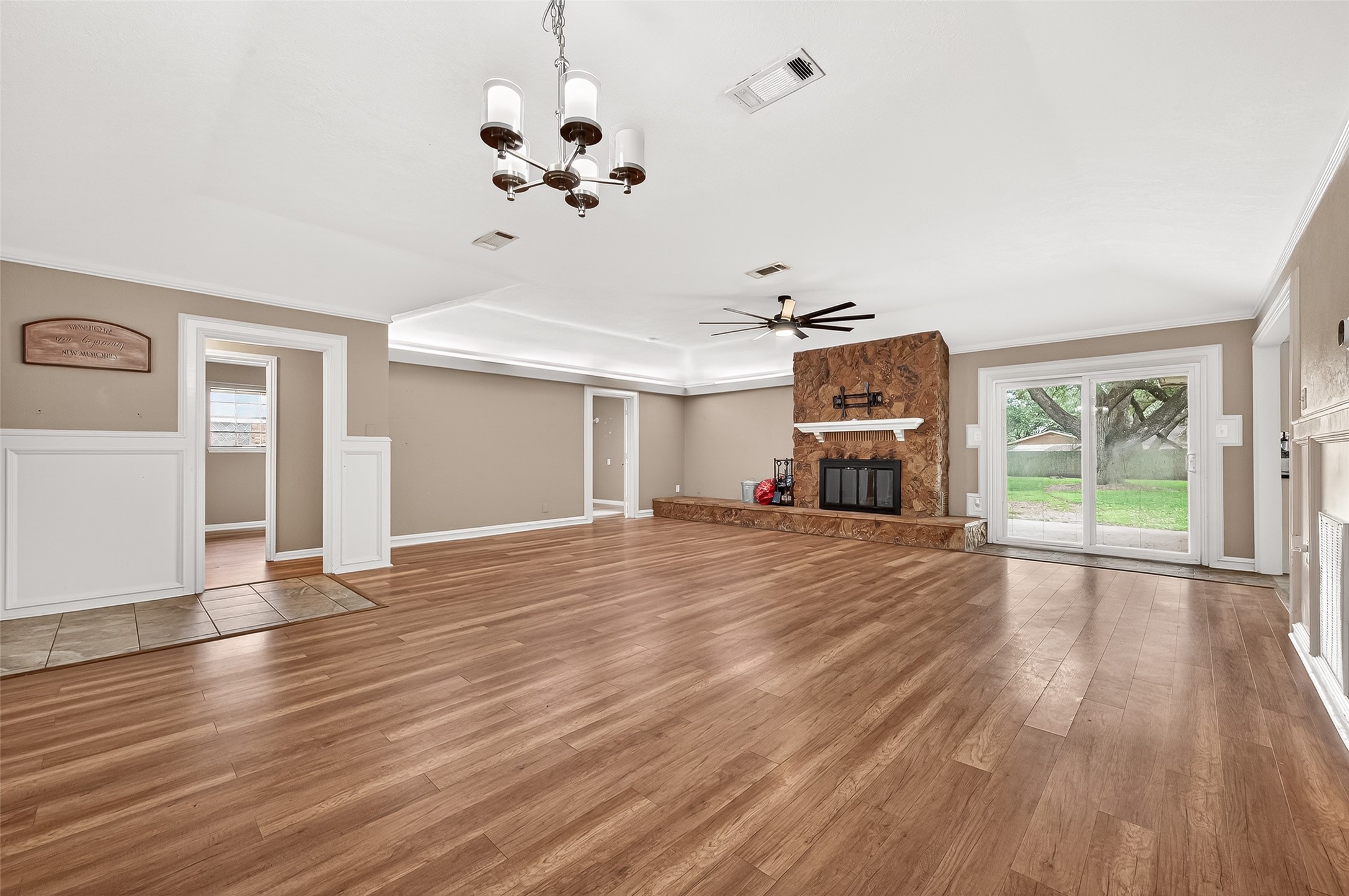 Spacious living room with elegant wood flooring and a cozy stone fireplace. Features a modern ceiling fan and a classic chandelier. Large glass doors offer a scenic view and natural light, leading to the backyard. Neutral wall tones provide a warm, inviting atmosphere.