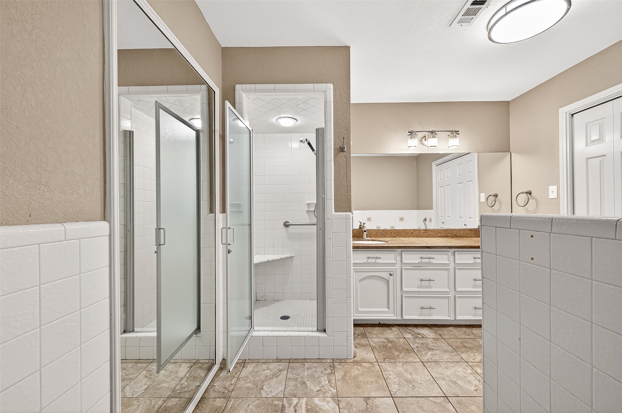 This bathroom features a spacious walk-in shower with glass doors, a large vanity with ample storage, and a bright, well-lit mirror. The neutral tones and tiled flooring create a clean, modern look.