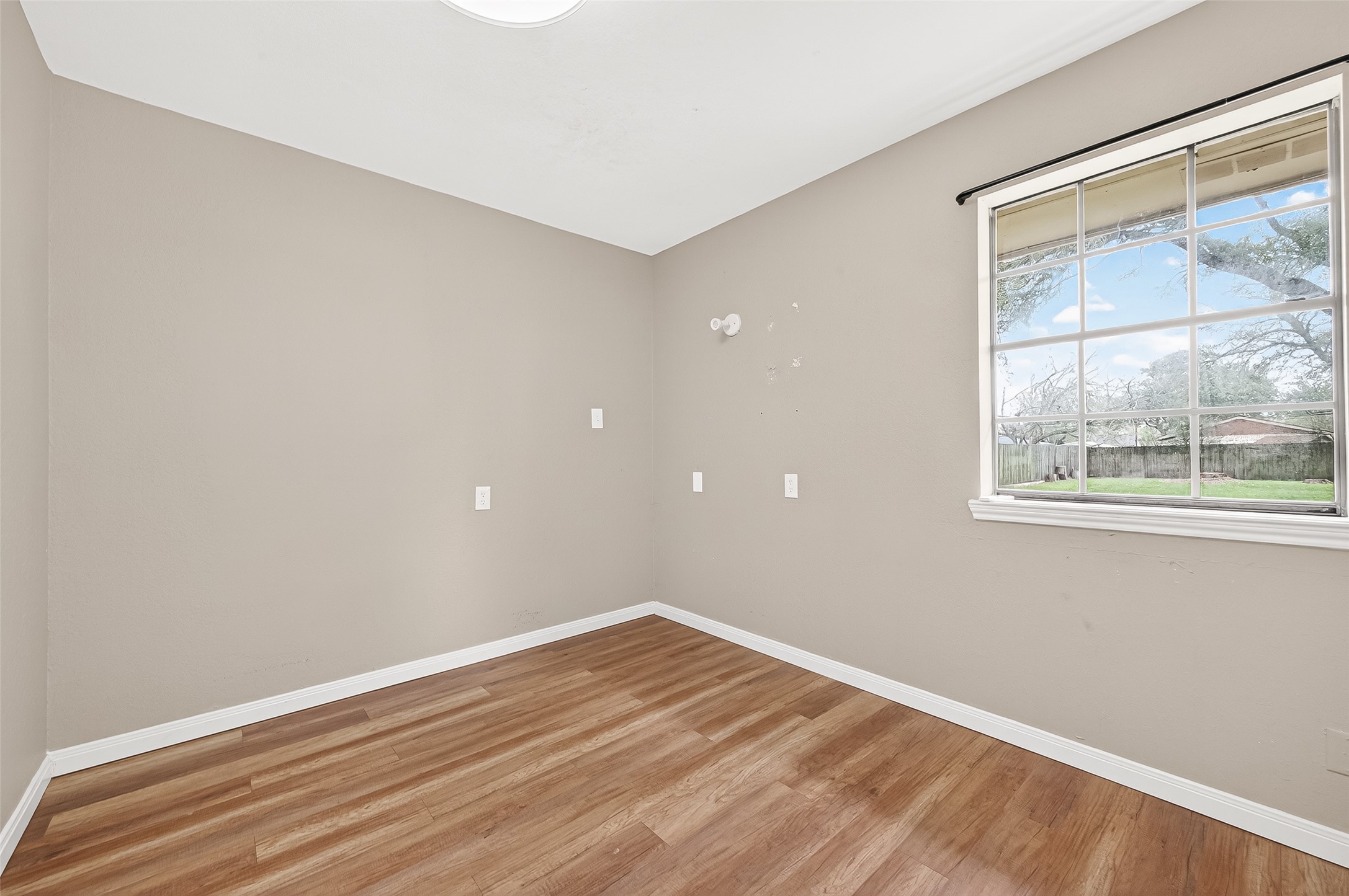 This room features neutral walls, wood flooring, and a large window offering plenty of natural light with a view of the backyard. Perfect for a small bedroom or home office.