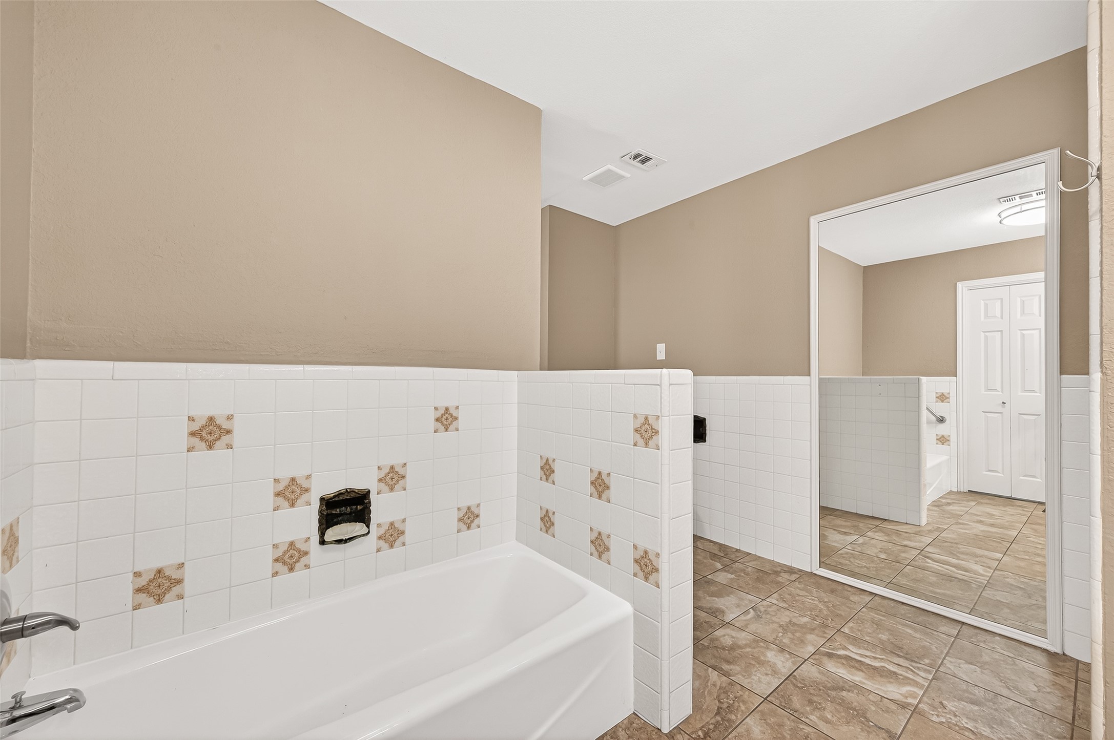 This bathroom features a spacious layout with a large bathtub, tiled walls with decorative accents, and a full-length mirror. The neutral color palette and tiled floor create a clean and modern look.