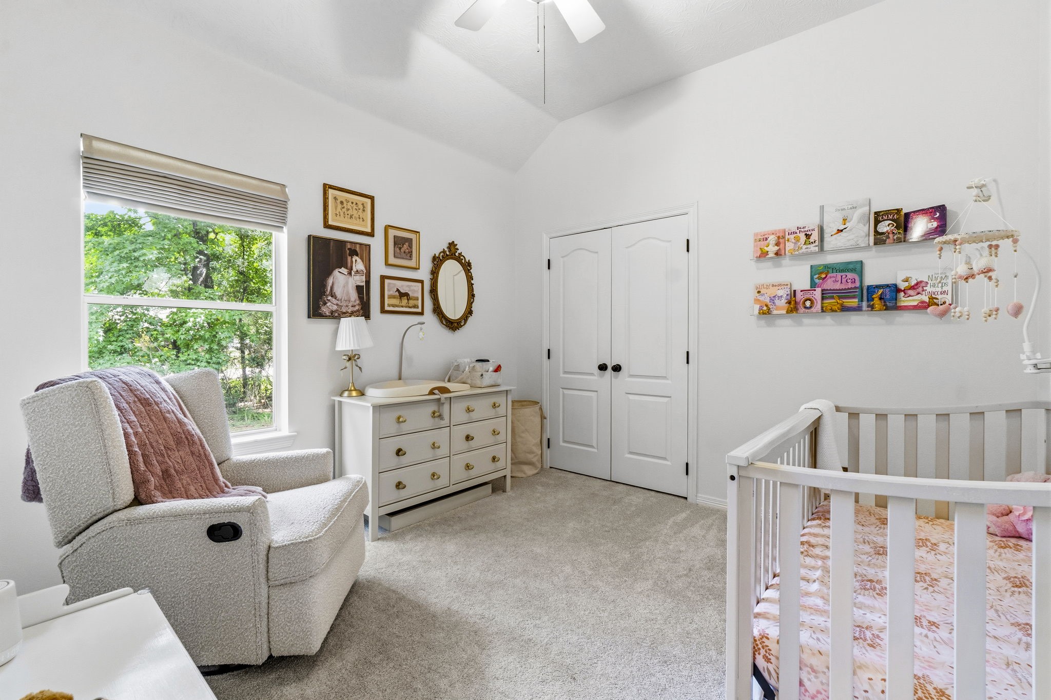 Charming nursery with soft carpeting, a cozy armchair, and a crib, enhanced by a large window bringing in natural light.