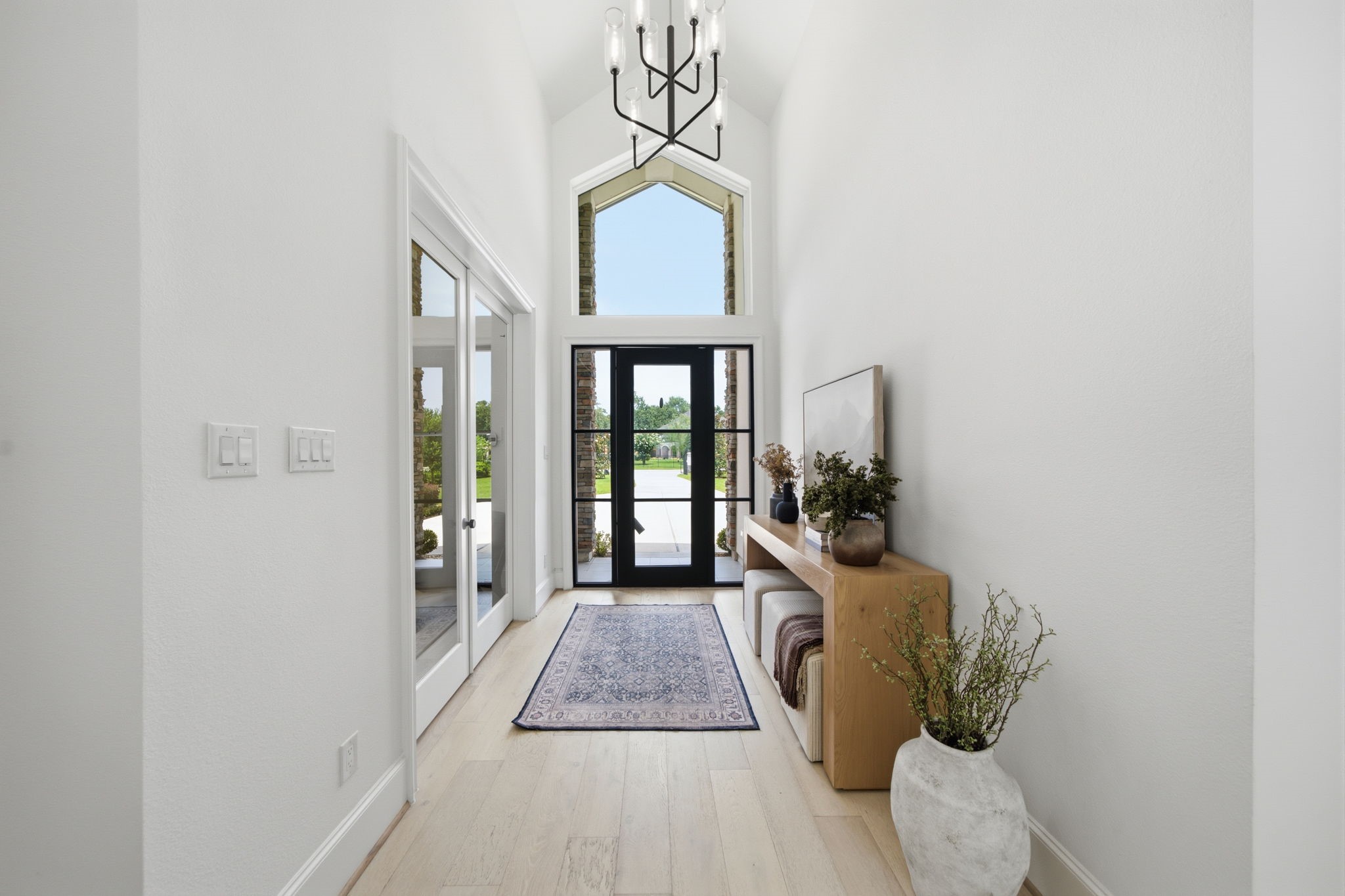 Bright and welcoming entryway featuring high ceilings, a modern chandelier, and large glass doors that flood the space with natural light, complemented by stylish decor and a neutral color palette. Bright and welcoming entryway featuring high ceilings, a modern chandelier, and large glass doors that flood the space with natural light, complemented by stylish decor and a neutral color palette.