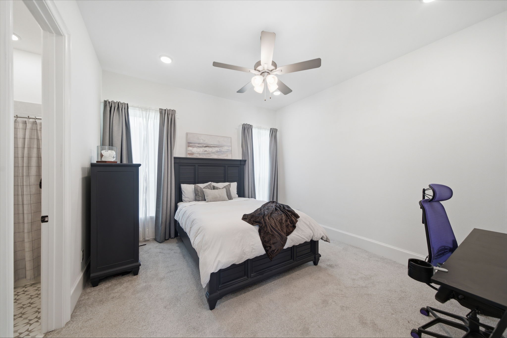 Spacious bedroom with cozy carpeting, featuring a stylish dark wood bed frame and matching furniture. Abundant natural light streams through the large windows, while a ceiling fan adds comfort. Adjacent en-suite bathroom enhances convenience.