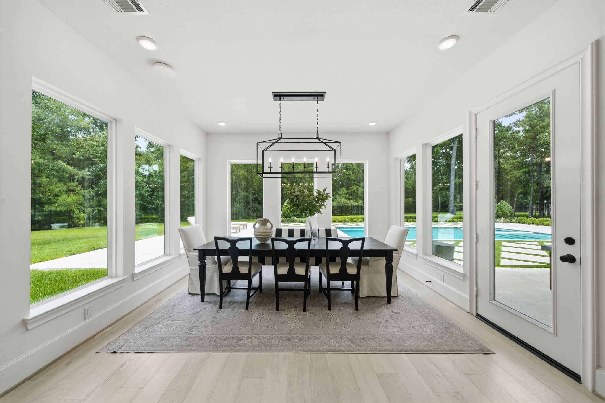 Bright and airy dining area with large windows showcasing a lush garden and sparkling pool view, featuring elegant lighting and a spacious table for gatherings.