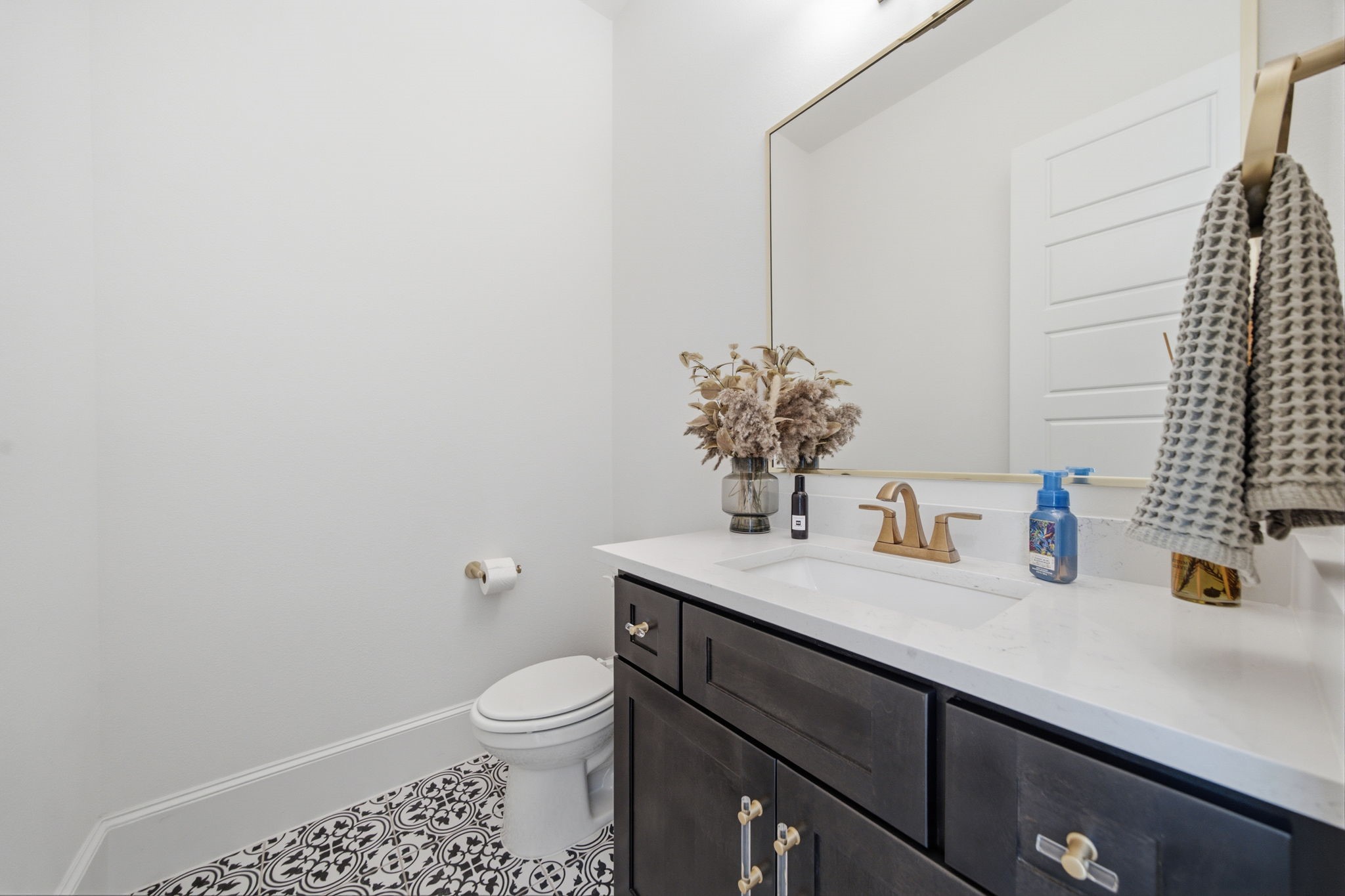 Elegant powder room featuring a sleek vanity with a quartz countertop, accented by a large mirror and stylish brass fixtures. The patterned tile floor adds a touch of sophistication.